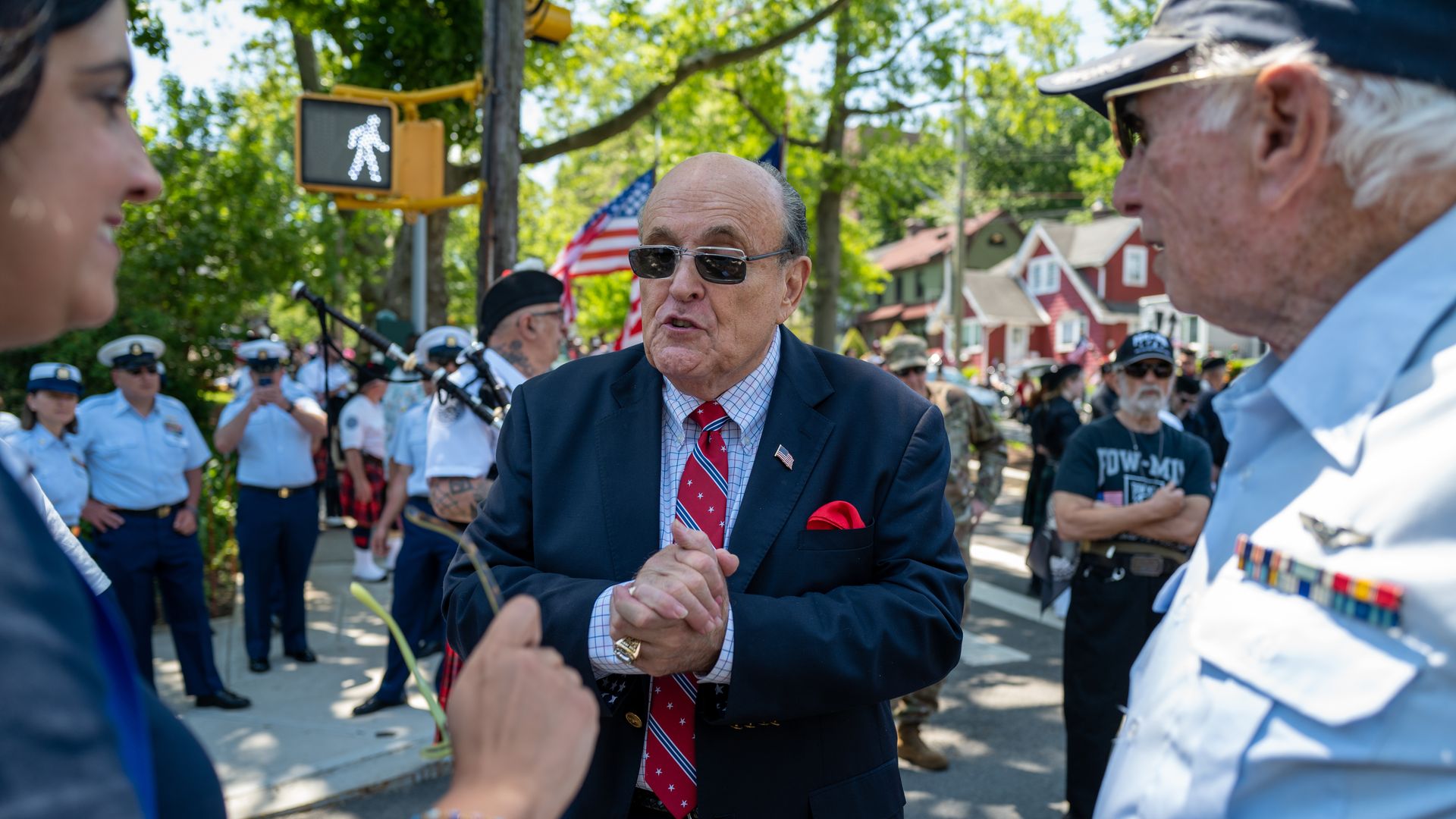 Photo of Rudy Giuliani wearing sunglasses and holding his hands clasped in front of him as he speaks to two people at an outdoor parade
