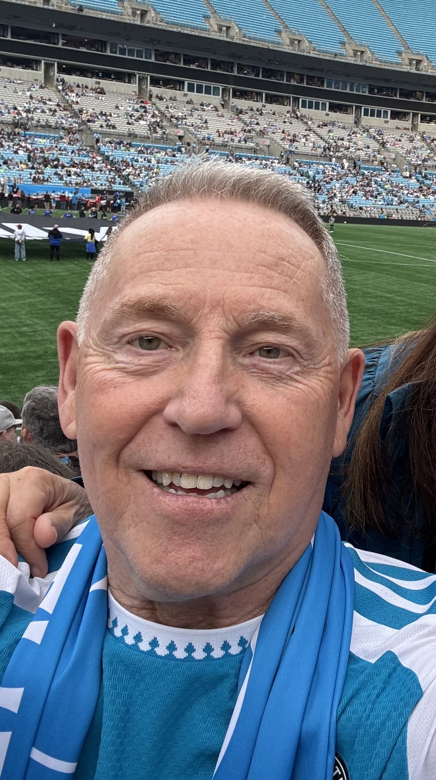 Close-up of a smiling man with short gray hair at a sports stadium; blue and white scarf around his neck, stands filled with spectators and a green field in the background.