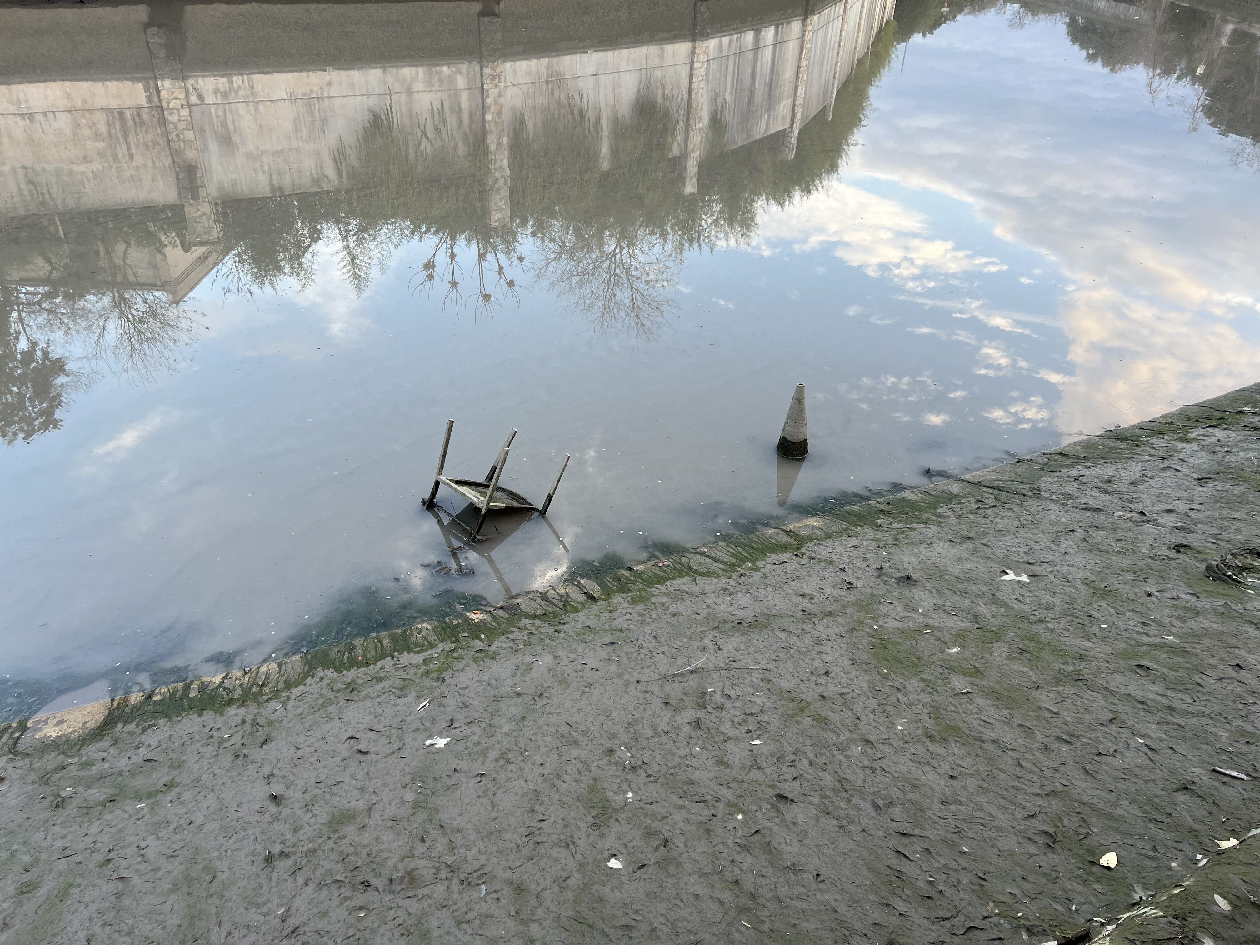 A muddy riverbank with a partially submerged upside-down chair and a cone in the water, reflecting the sky and surrounding trees and concrete wall.