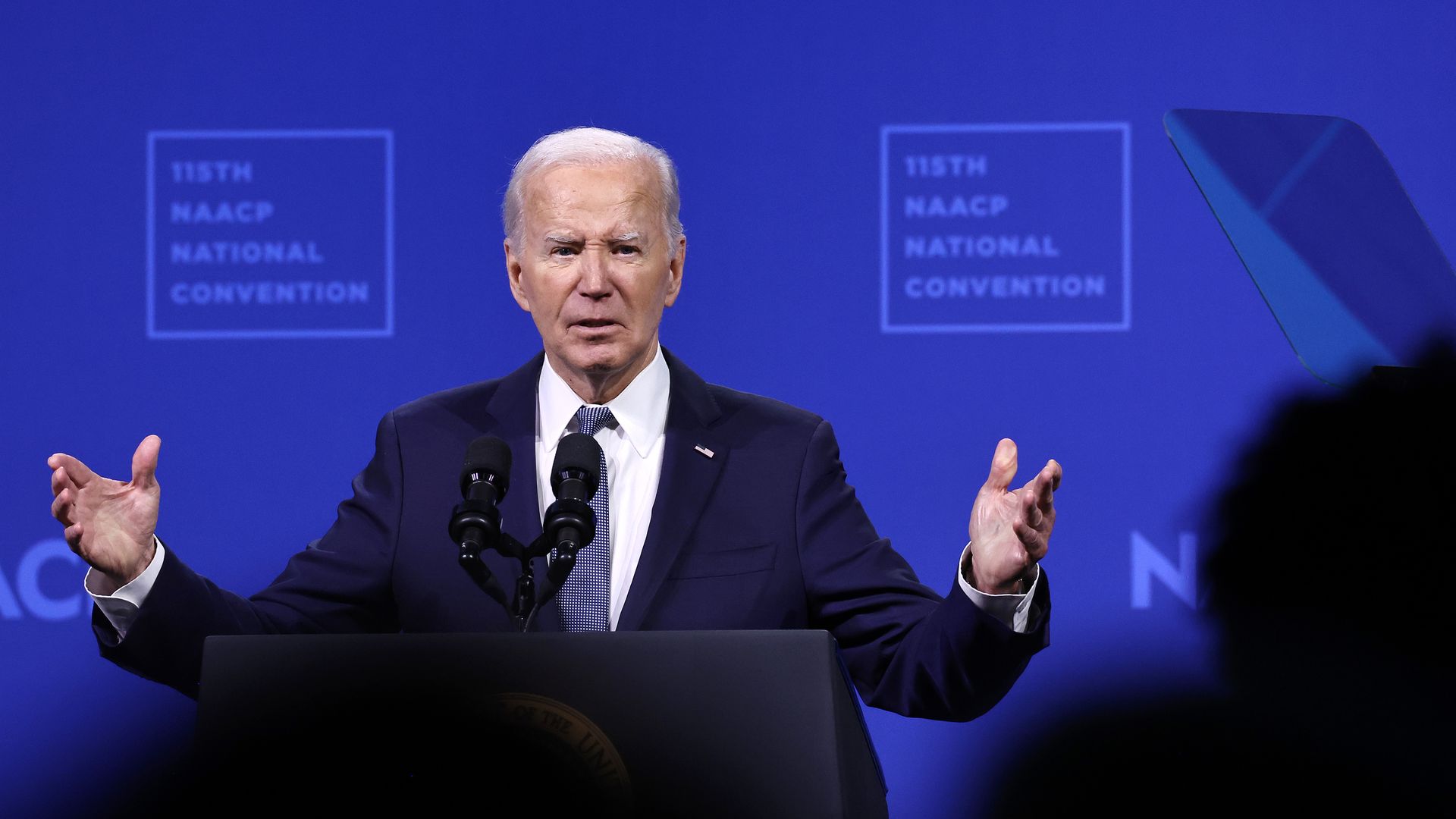 Biden speaks into a microphone at a lectern. His hands are outstretched in front of him. Behind him is a blue wall that says 115th NAACP National Convention. 