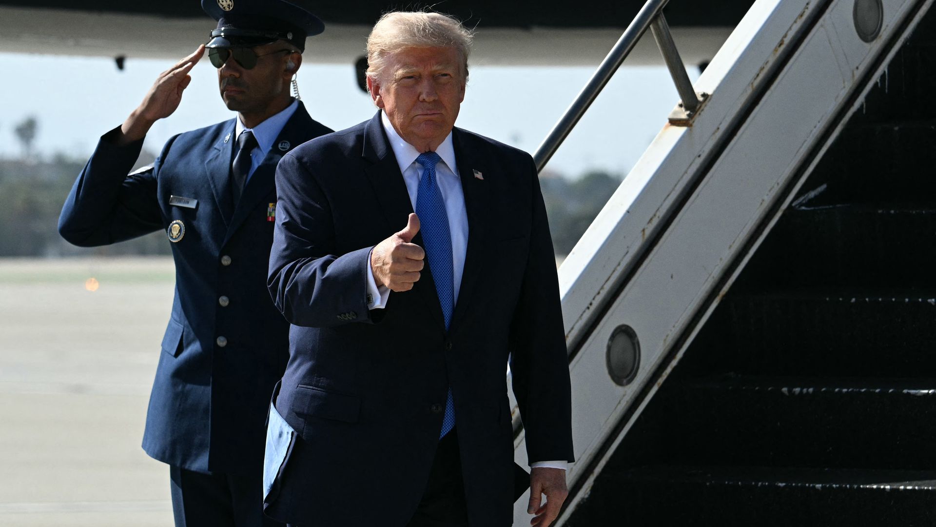 President Trump, in a dark suit and blue tie steps from aircraft stairs, giving a thumbs-up; a uniformed security officer salutes beside him on the tarmac.