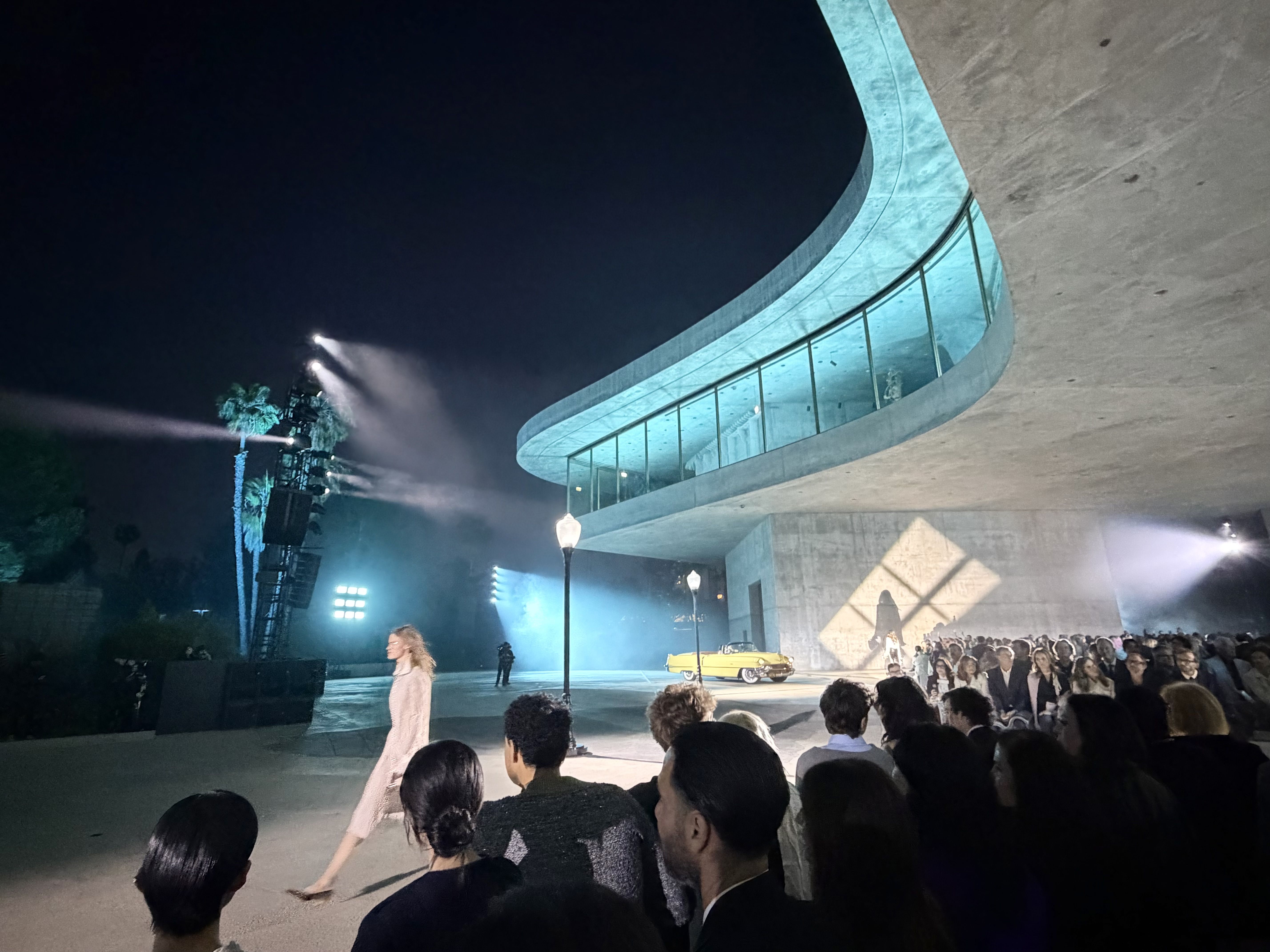 Nighttime outdoor event showing a curved turquoise-lit concrete walkway with glass railing, a seated crowd to the right, stage lights, and a yellow vintage car in the background.