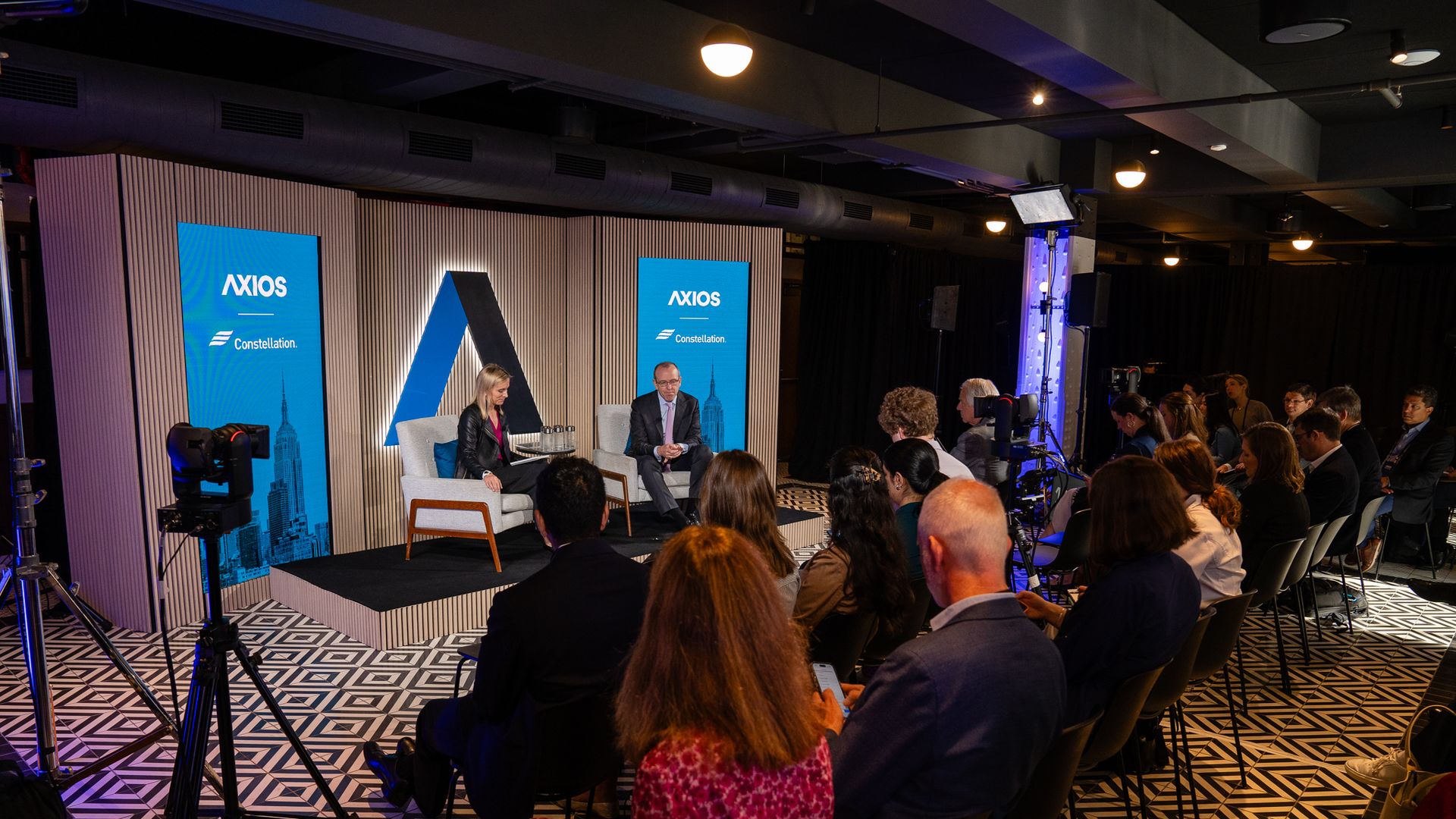 Audience seated facing two speakers on stage at an Axios event with blue banners, geometric black-and-white flooring, and professional recording equipment.