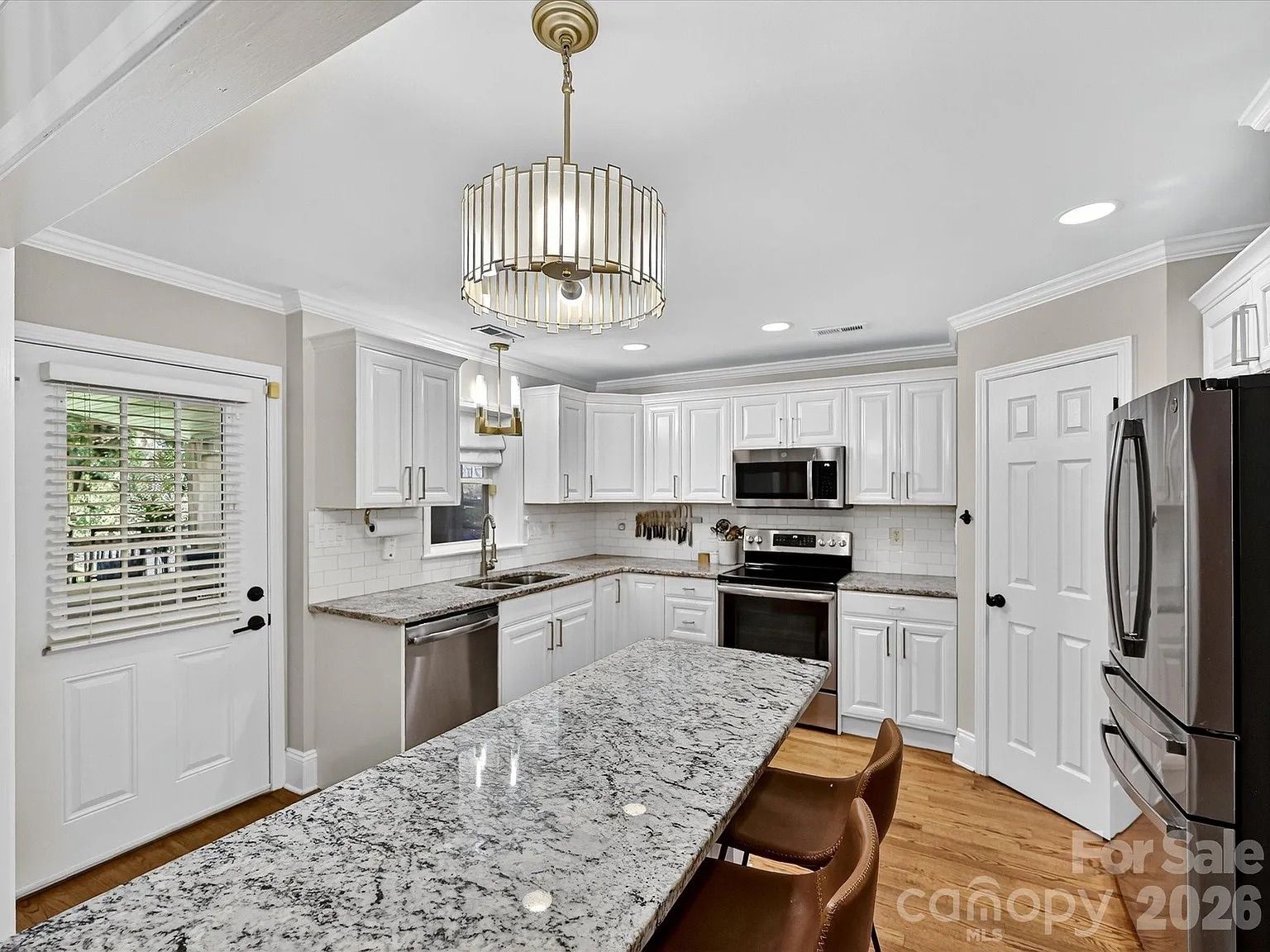 Modern kitchen with white cabinets, granite countertops, stainless steel appliances, hardwood floors, two brown chairs at the island, and a decorative pendant light above.