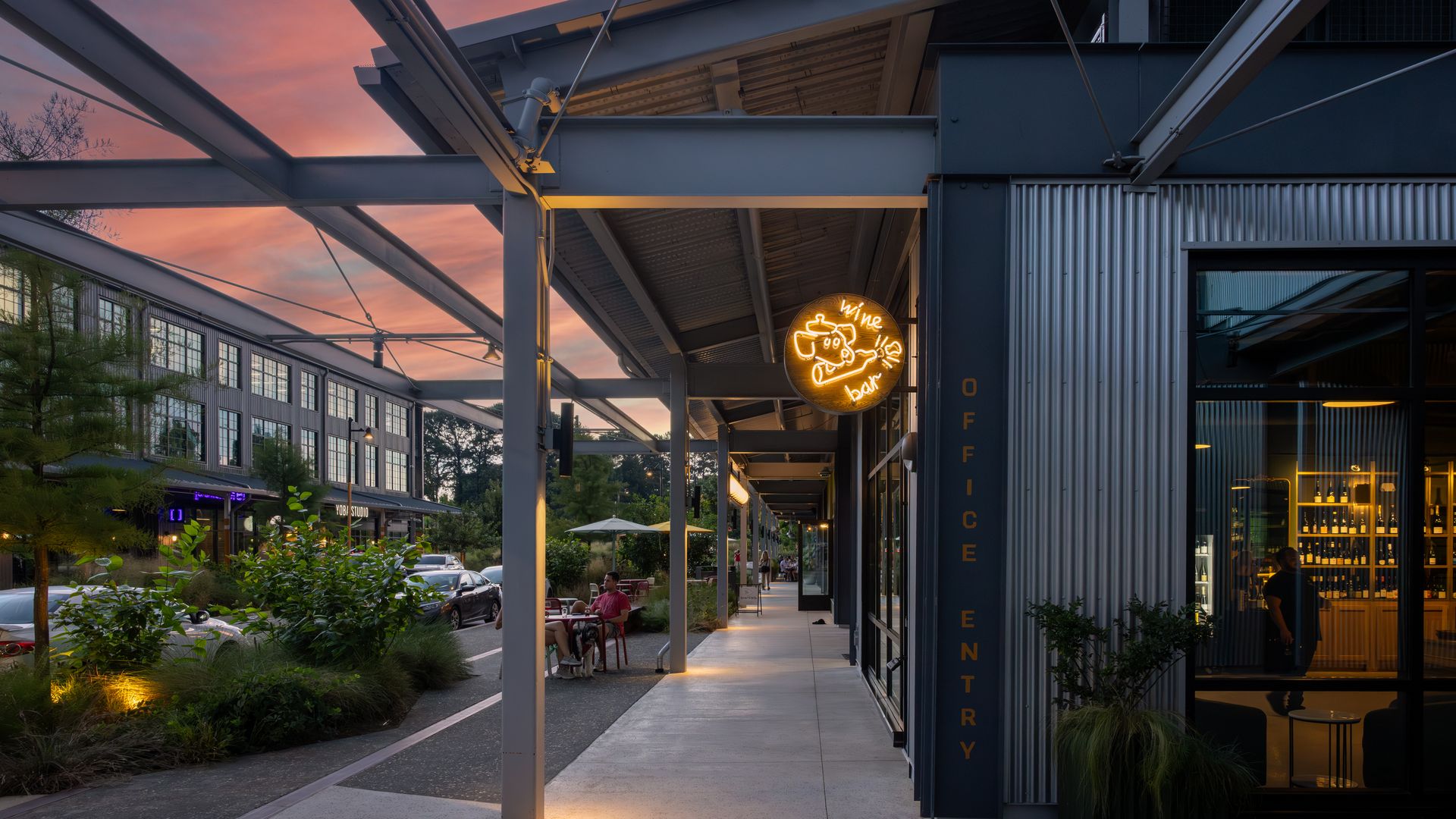 Outdoor walkway at dusk with modern buildings, illuminated neon "wine bar" sign, people seated at tables, and purple-pink sky with clouds above.