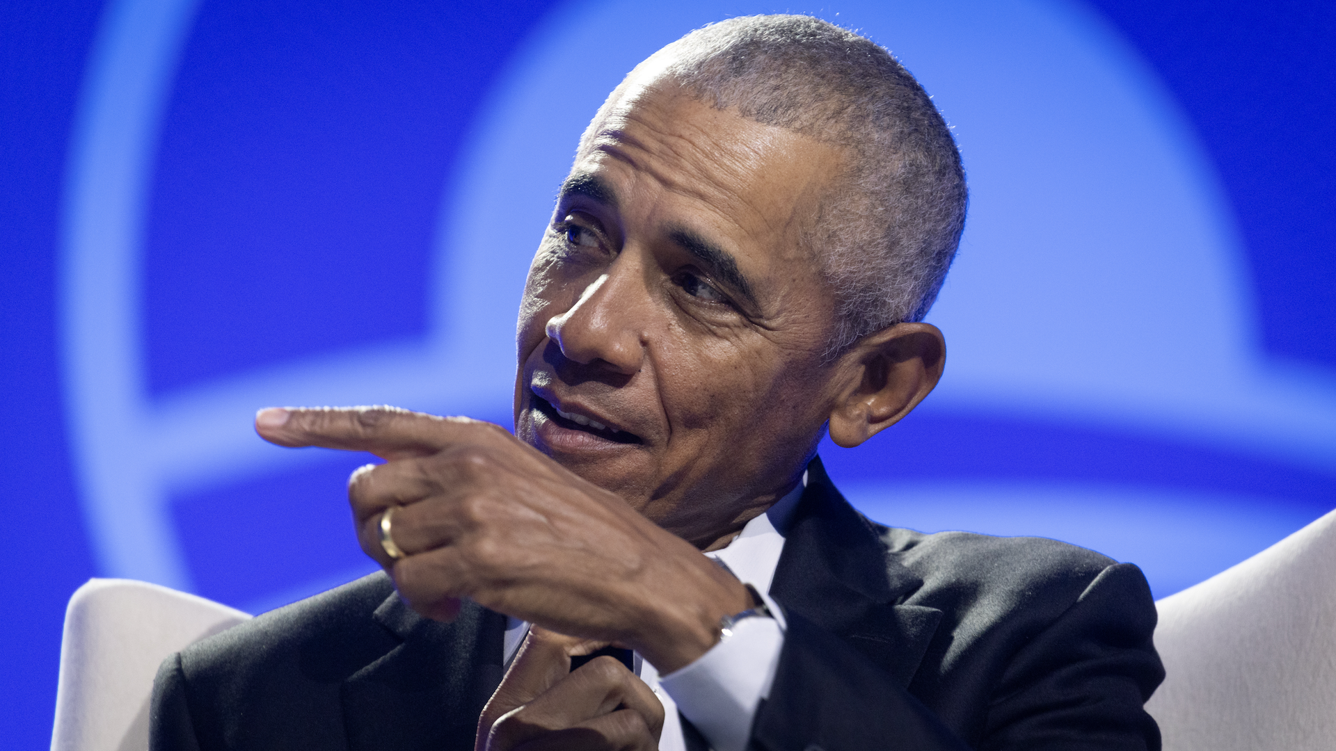 Barack Obama, the first Black US president wearing closely cropped, dark graying hair, points with his left hand, showing a gold ring on his wedding ring finger, while sitting in a cream chair and wearing a black suit jacket and white shirt, before a blue background.