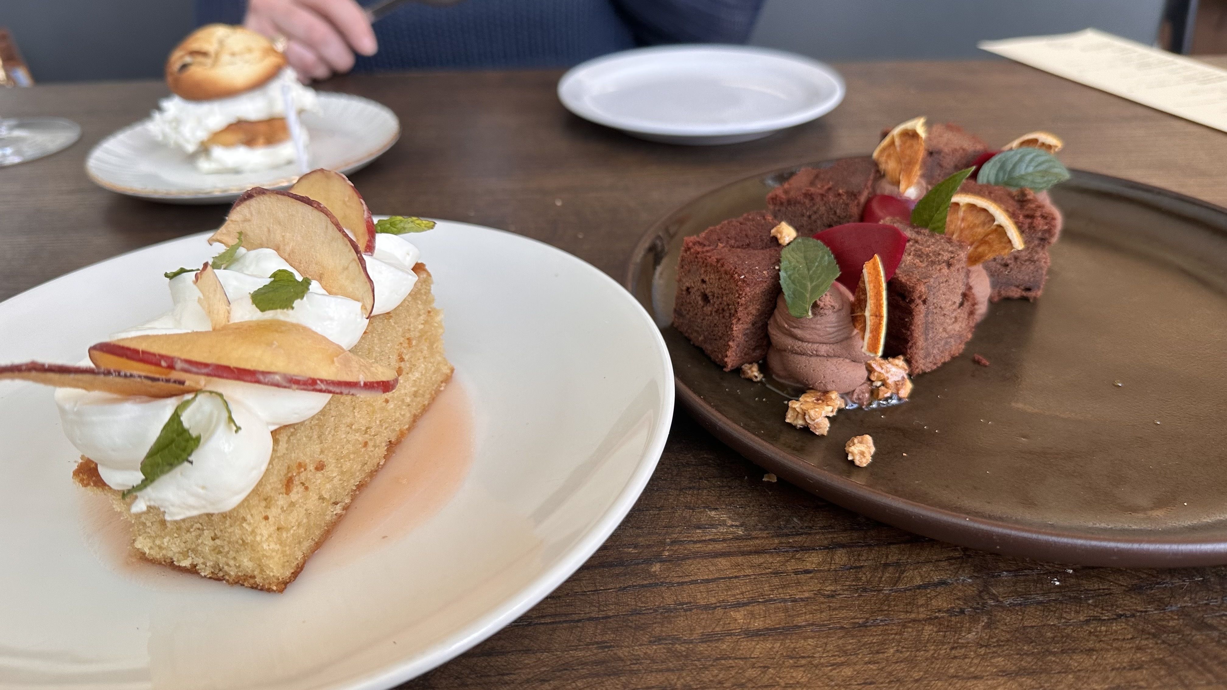 Two desserts on a wooden table: a whipped-cream topped sponge cake with apple slices on a white plate, and a chocolate cake with mousse, orange slices, and mint on a dark tray; a blurred person in a blue top sits in the background.