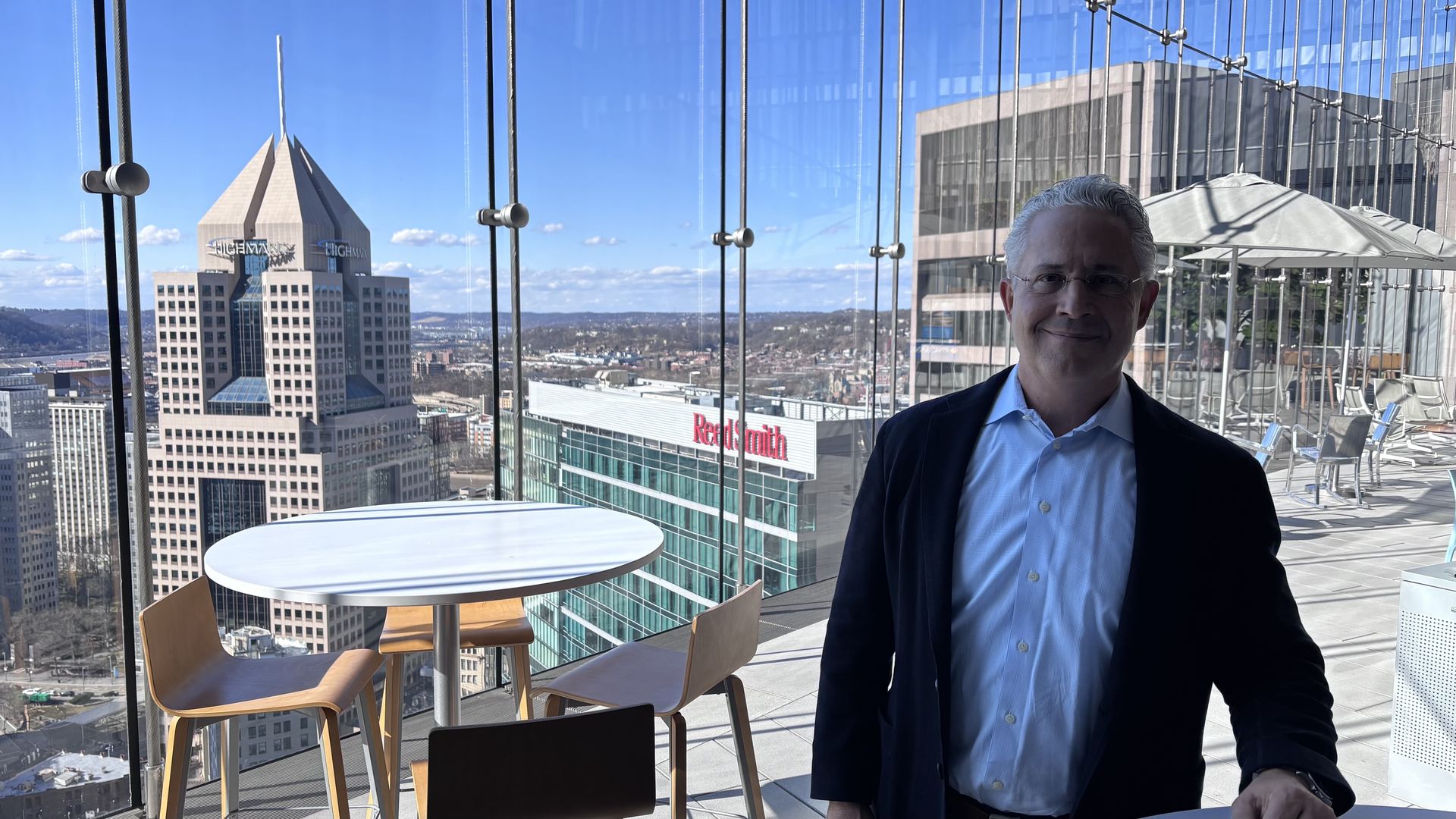 Man in glasses and dark jacket stands on a glass-walled balcony with a round white table and wooden chairs, overlooking a city skyline including the Highmark building and Reed Smith office.