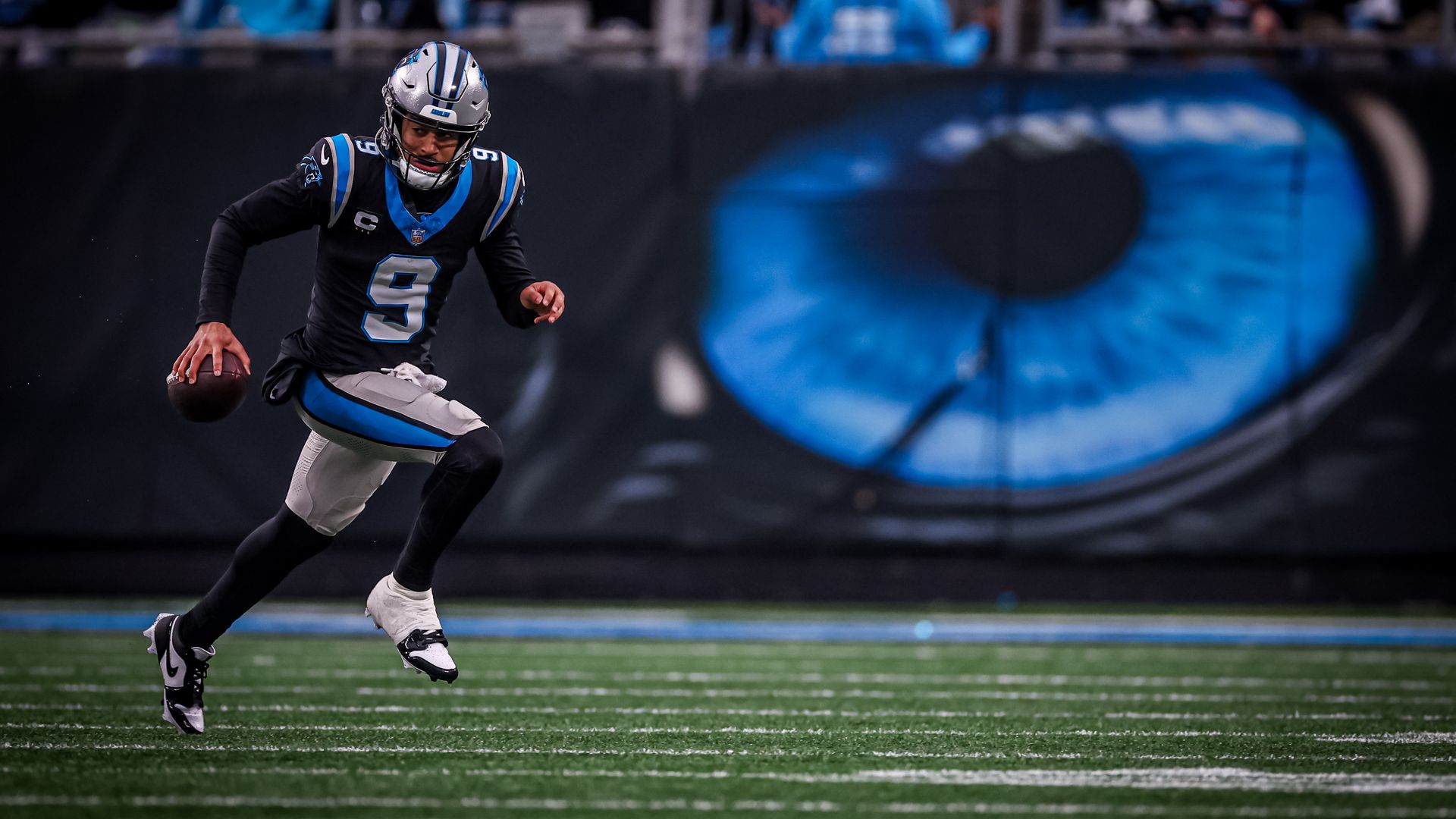 Carolina Panthers quarterback Bryce Young runs the ball on the field, with a large blue cat eye painted behind him.