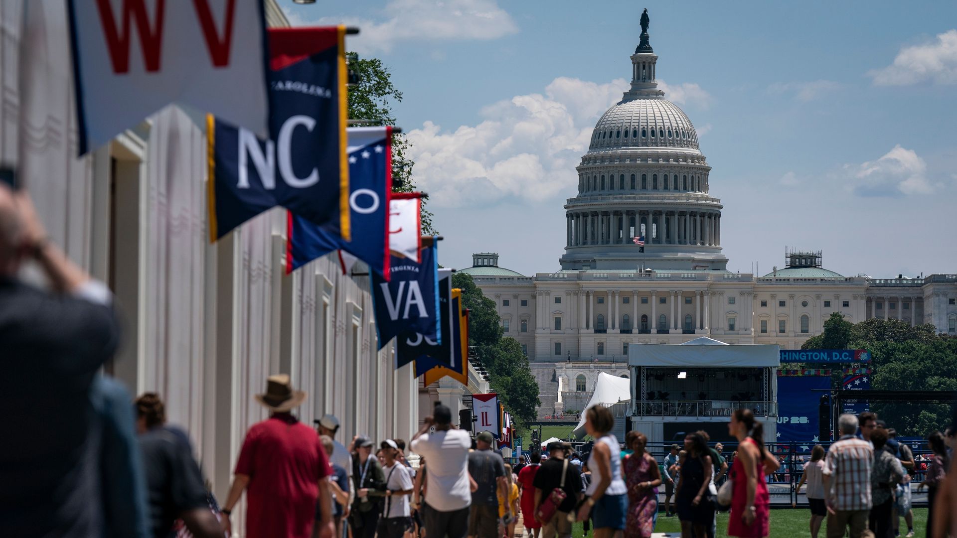 Crowd walks along a parade route with colorful flags lining the left, as the U.S. Capitol dome rises under a blue sky on a sunny day.