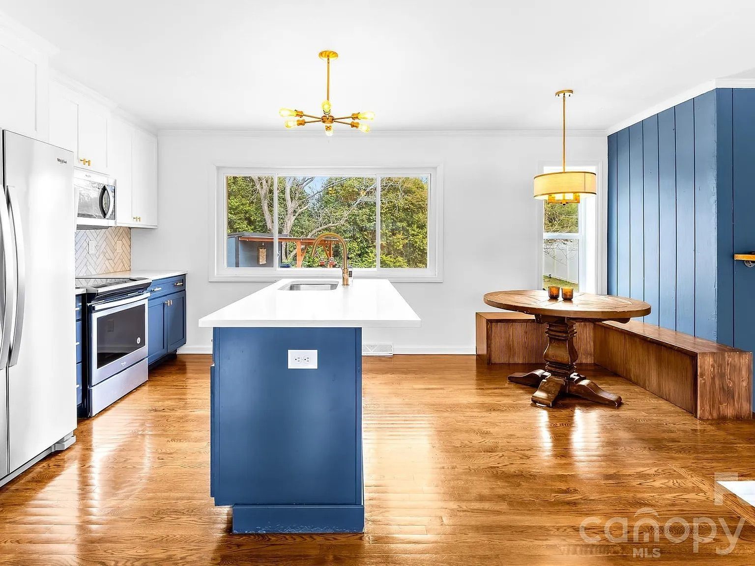 Bright kitchen with blue cabinets, white countertops, hardwood floors, a large window showing greenery outside, a round wooden dining table with a built-in bench, and modern gold light fixtures.