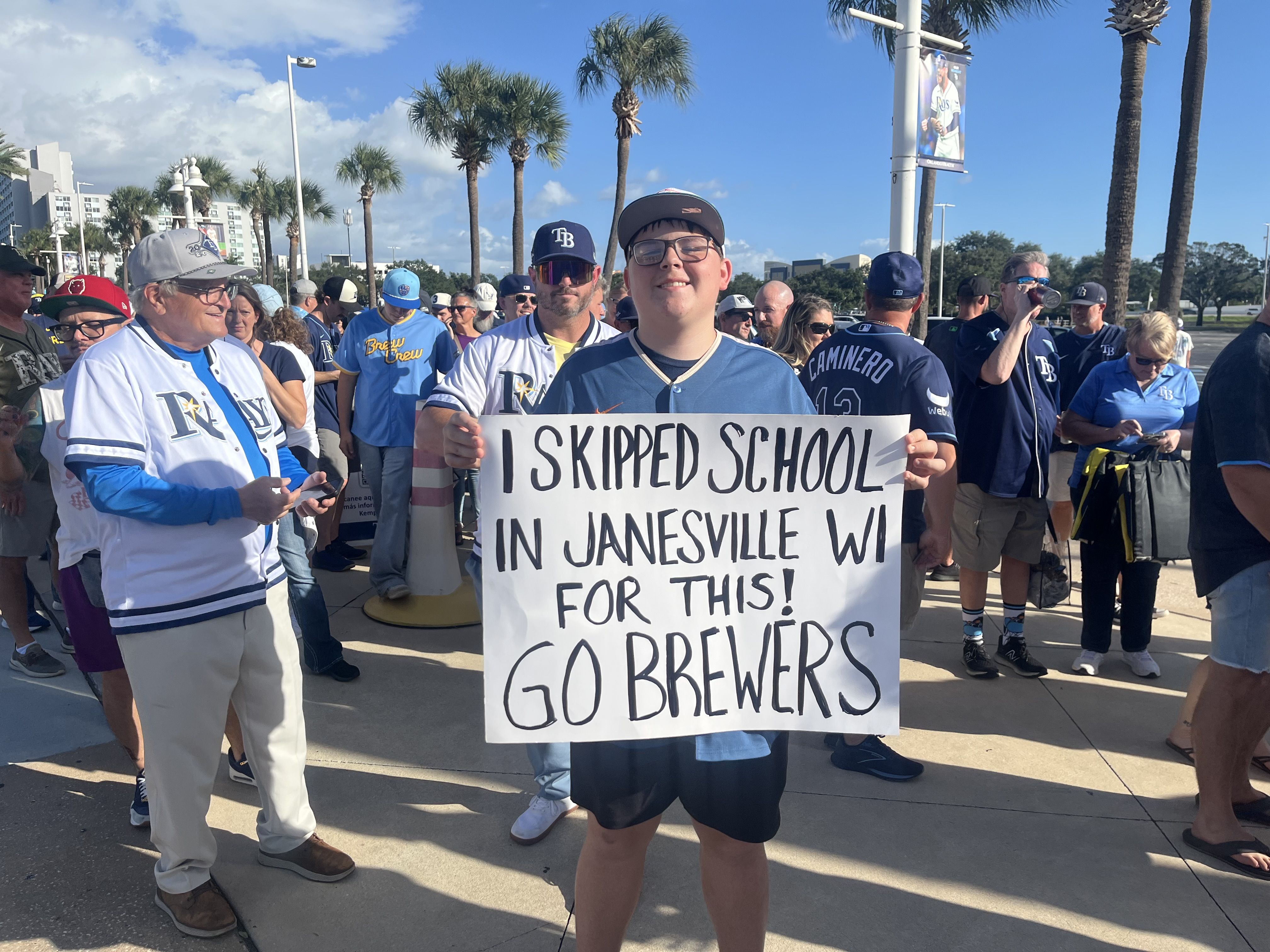 Outdoor crowd of baseball fans in Rays and Brewers gear under palm trees on a sunny day; the center person holds a large sign reading, "I SKIPPED SCHOOL IN JANESVILLE WI FOR THIS! GO BREWERS".
