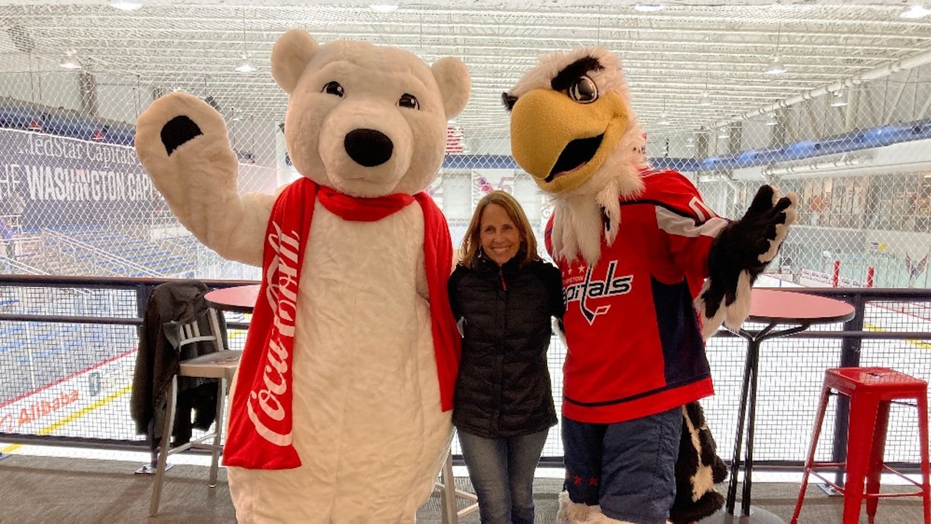 A woman poses between the Coca-Cola polar bear mascot and the Washington Capitals mascot at an indoor ice rink.