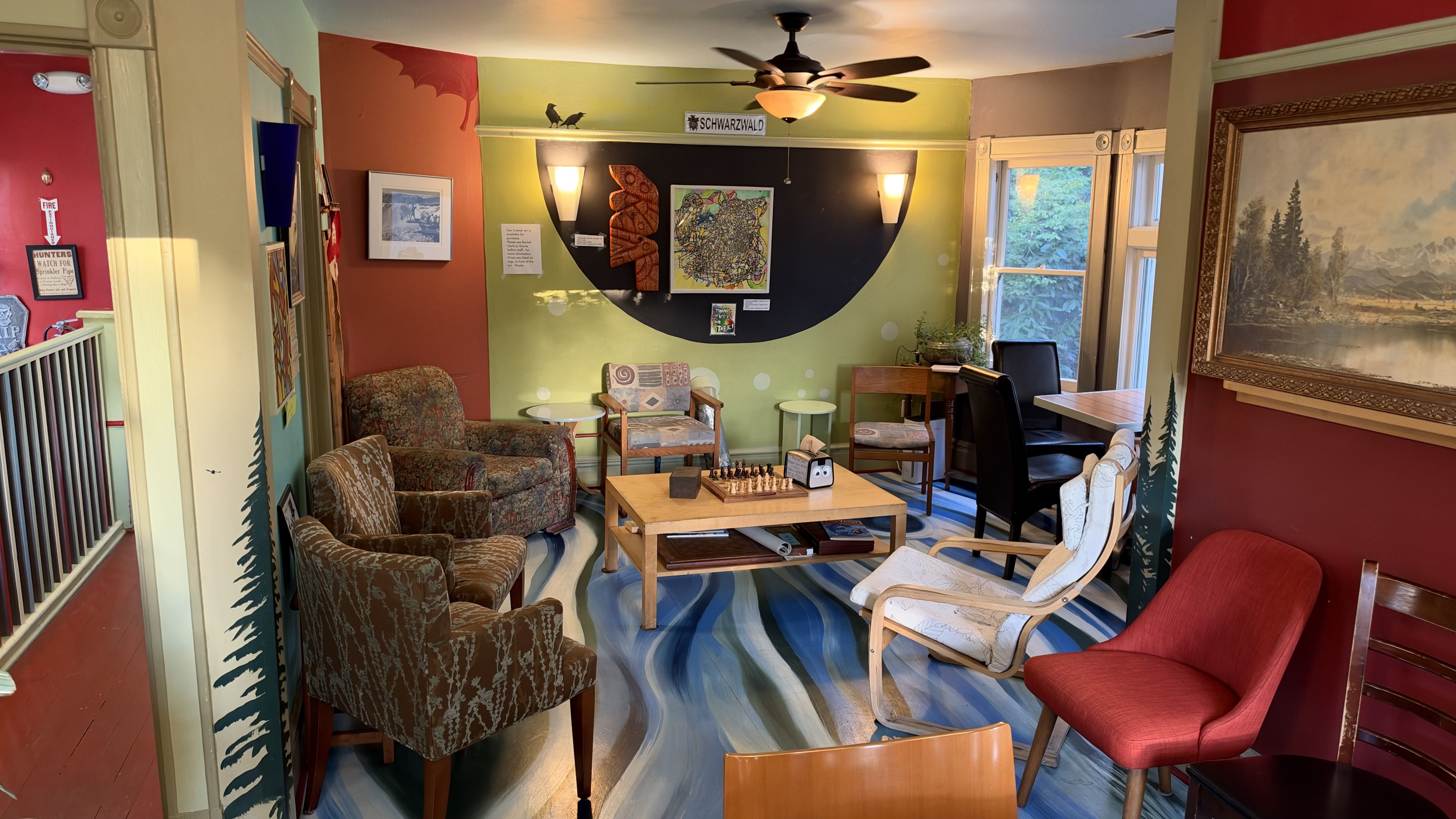 Colorful living room with red walls, a large mural featuring a black semicircle, patterned armchairs, wooden chairs, a chessboard on the coffee table, ceiling fan, and a view of trees through windows.