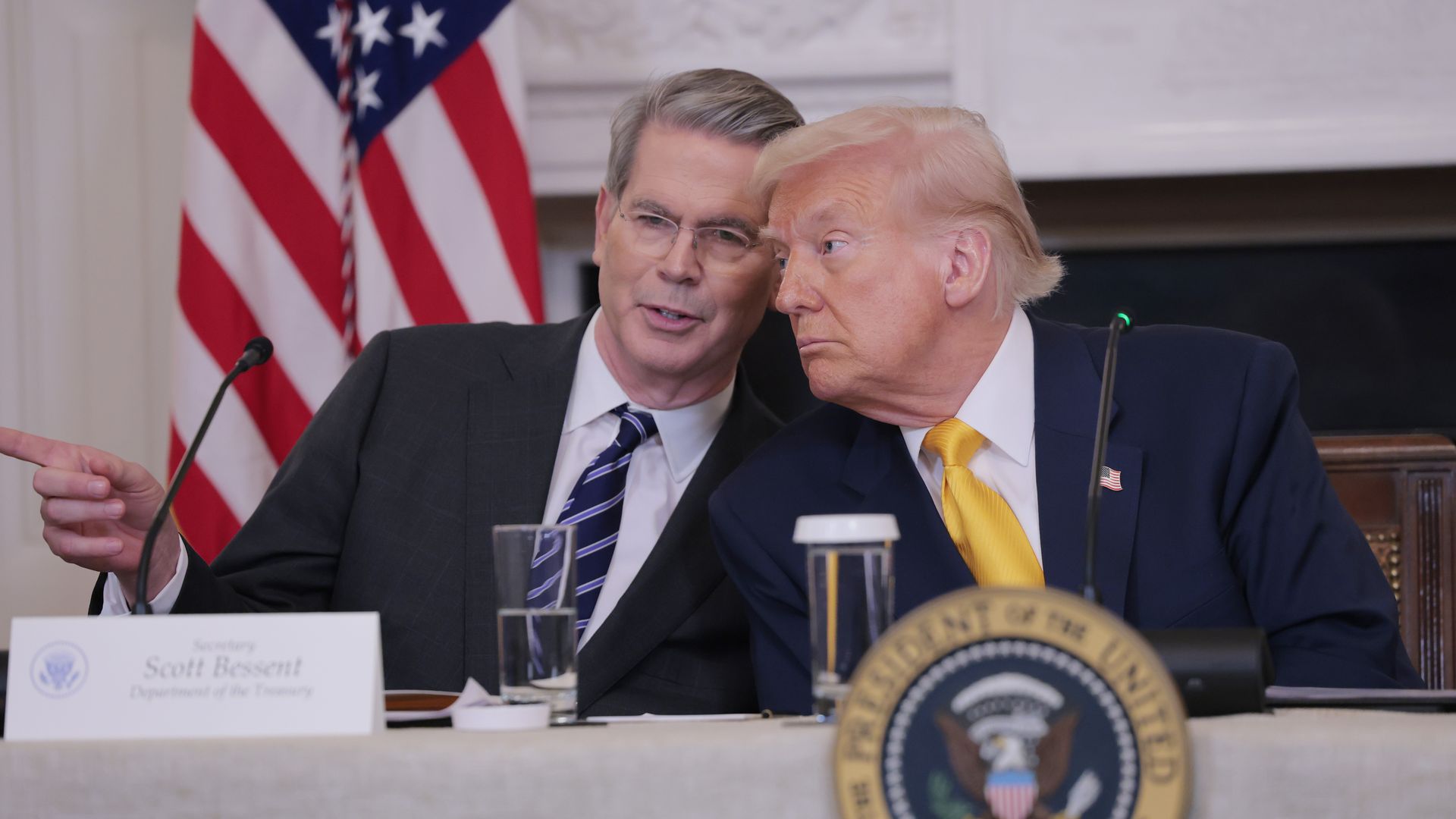 Treasury Secretary Scott Bessent and President Trump in dark suits sit at a conference table; one leans in toward the other as they speak. An American flag stands behind them and the presidential seal is visible on the desk.