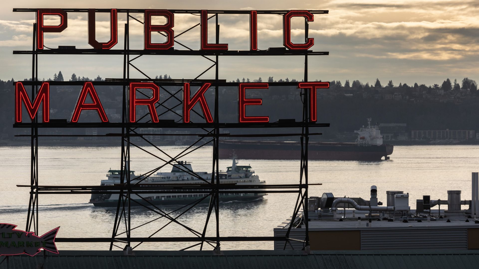 A photo of the Public Market sign at Pike Place Market with a sunset behind and a ferry crossing Elliott Bay. 