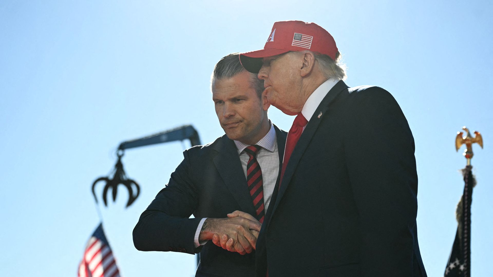President Trump — wearing a dark suit, a white collared shirt, a red tie and a red hat — shakes hands with Pete Hegseth, who is wearing a dark jacket, a white collared shirt and a striped red and dark tie.