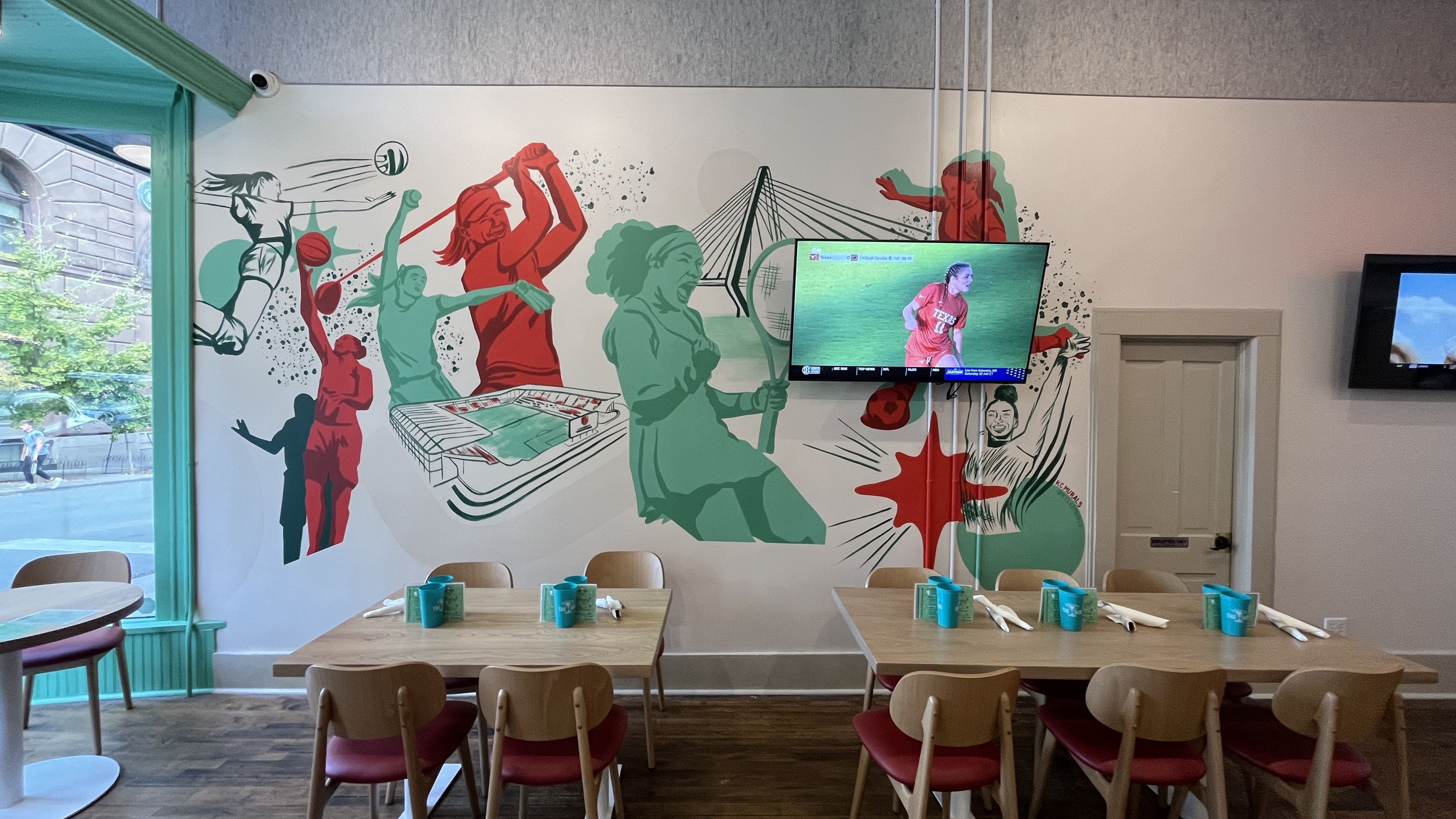 Interior of a The Dub with wooden tables and chairs, teal mugs, and a colorful mural on the wall depicting various sports like volleyball, tennis, and golf in red and green tones.