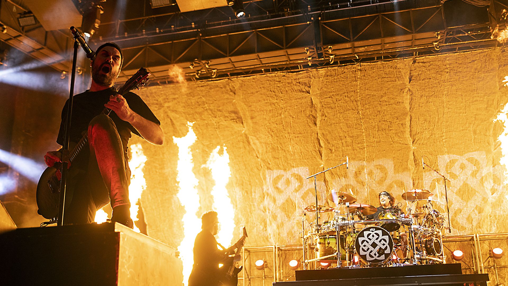 A guitarist singing while fire shoots out on stage behind a drummer in the back