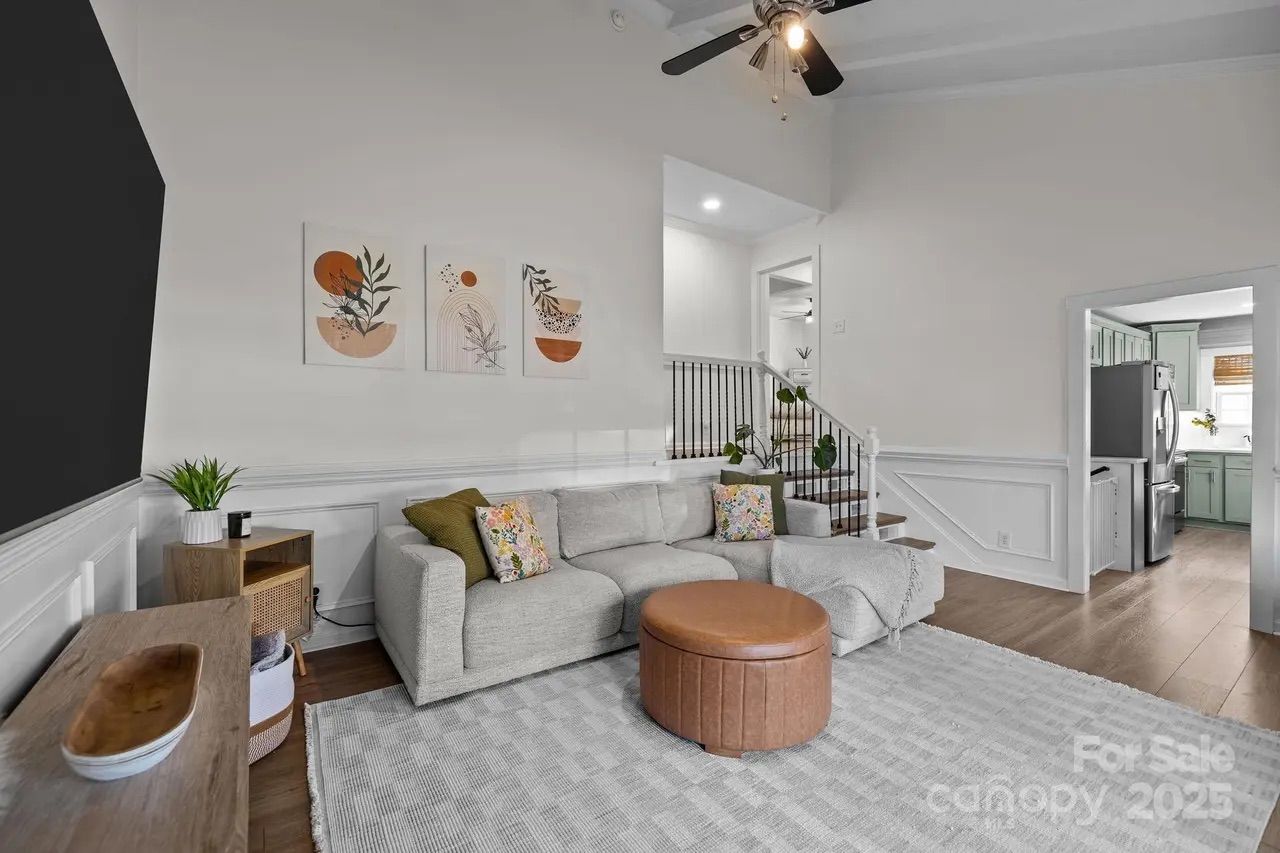Living room with gray sectional couch, brown round ottoman, light gray patterned rug, wooden furniture, three abstract wall art pieces, white walls, and ceiling fan. Kitchen visible in background.