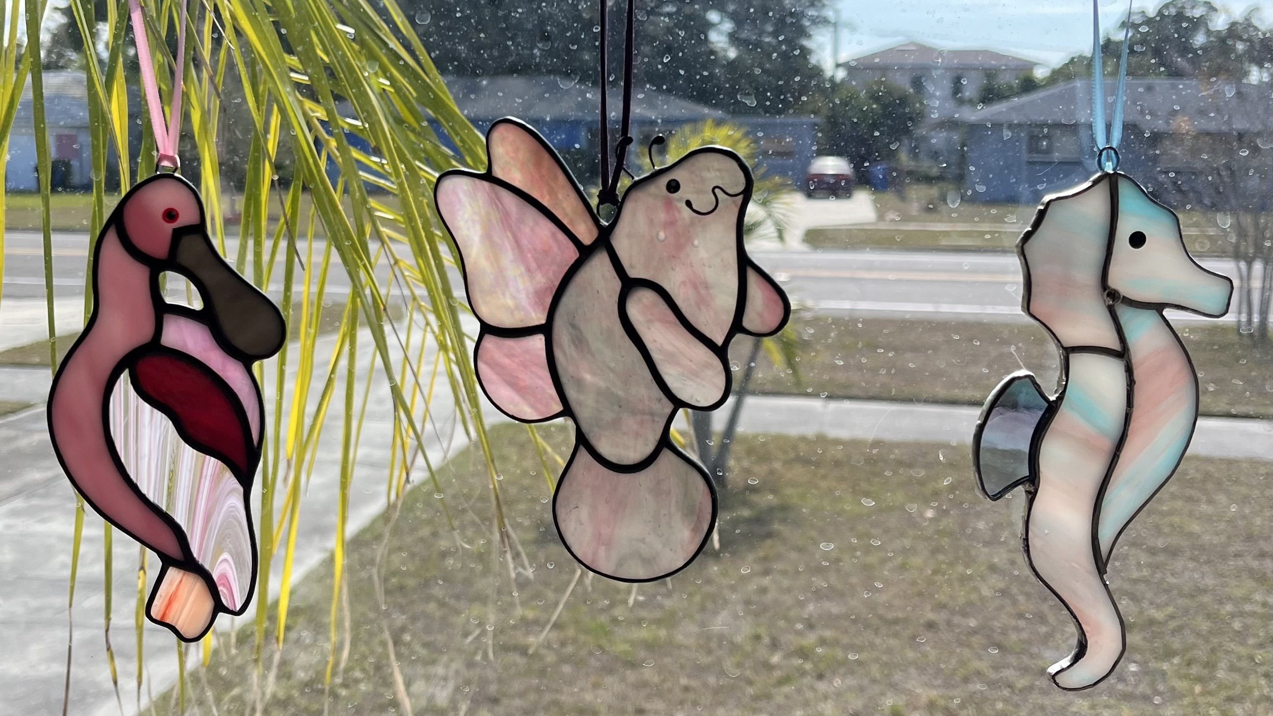 Three colorful stained glass window decorations shaped like a pink flamingo, a smiling manatee with fairy wings, and a pastel blue-pink seahorse hanging on a sunny window with a street and houses outside.