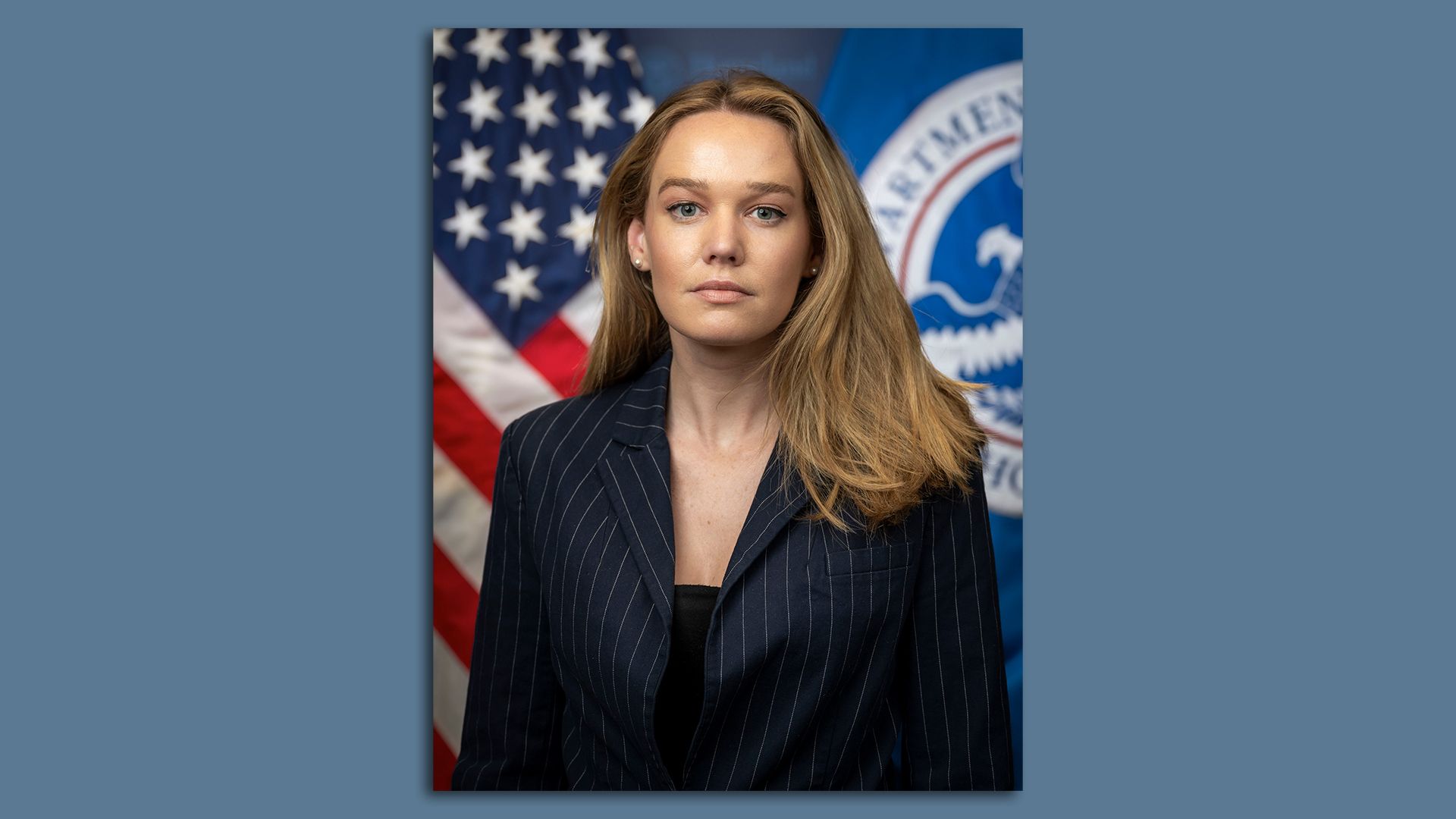 Portrait of Tricia McLaughlin, a woman with long blonde hair wearing a dark pinstripe blazer, standing in front of U.S. flags, including the American flag and a blue Department of Homeland Security flag.