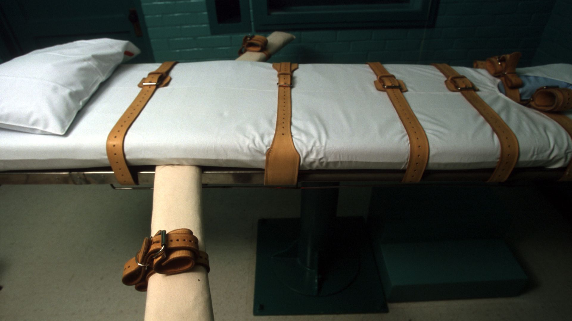 White restrained bed with multiple brown leather straps in a dimly lit room with teal brick walls and a pillow at one end.