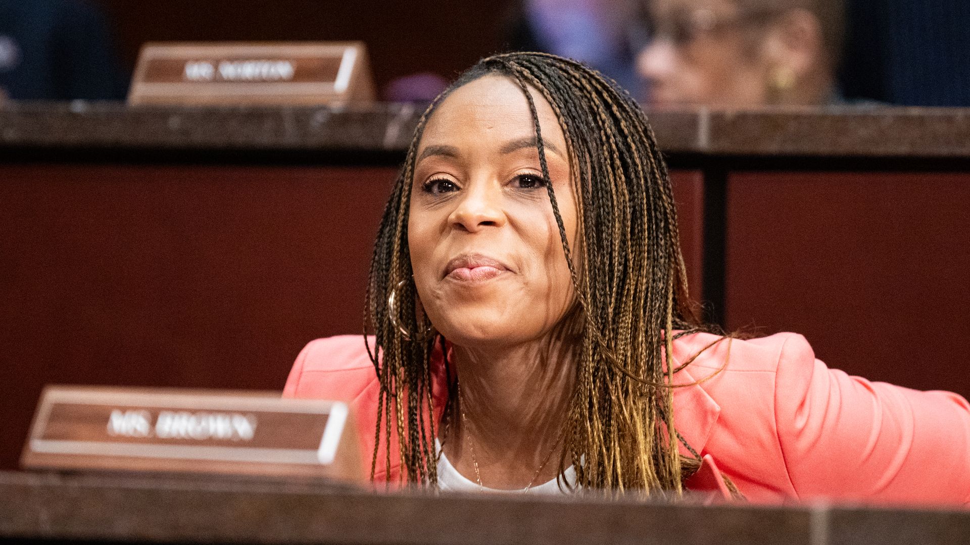 Photo of black woman with braids and a salmon-colored suit smirking at camera. Name sign at her seat says "Ms. Brown" 