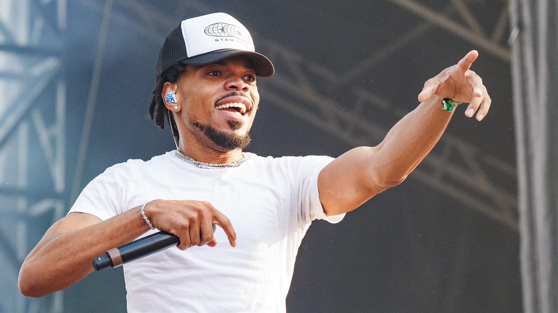Smiling male performer in a white t-shirt and Star Line black-and-white cap holds a microphone, reaching out with his right hand on a stage with dark background and metal truss.