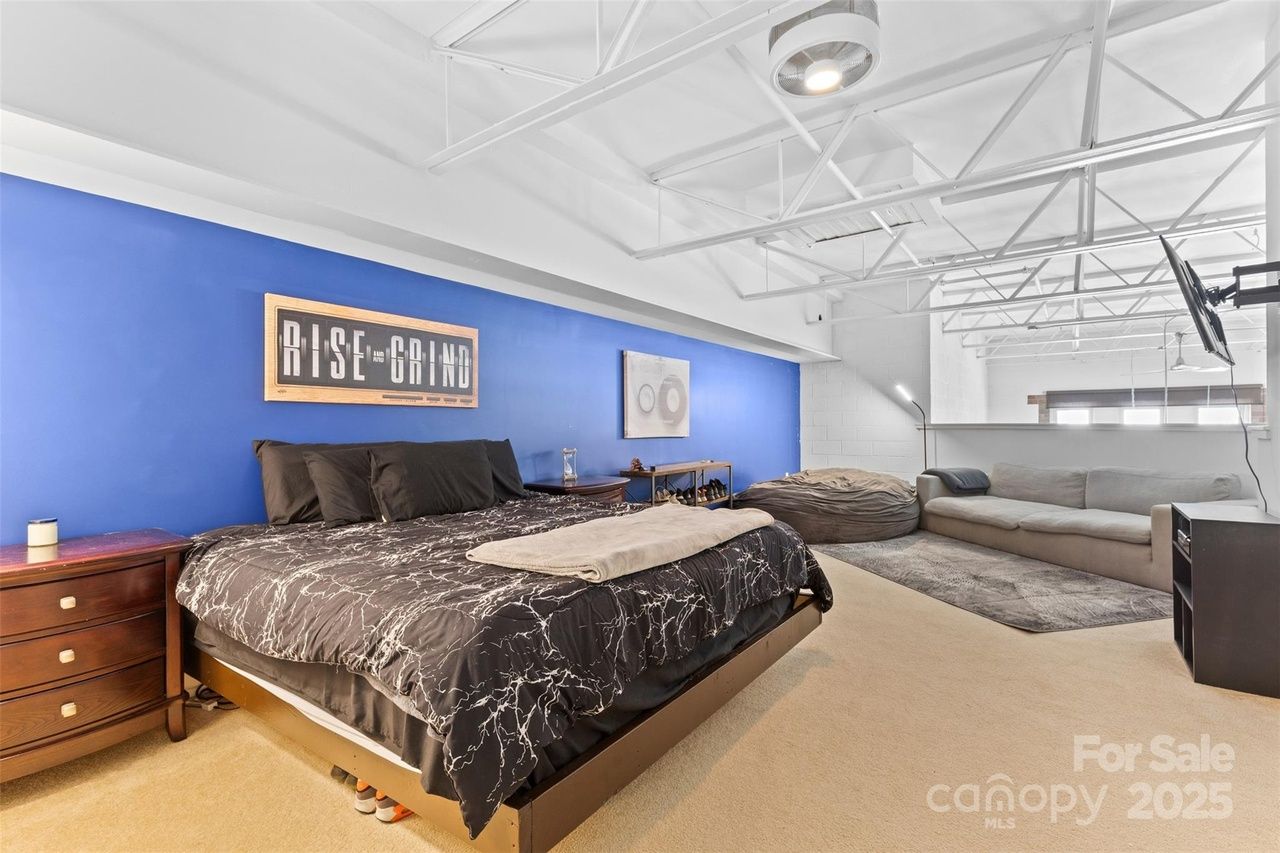 Modern bedroom with black bedding on platform bed, blue accent wall with "Rise and Grind" sign, wooden nightstands, beige carpet, bean bag, gray sofa, mounted TV, and exposed white ceiling beams.