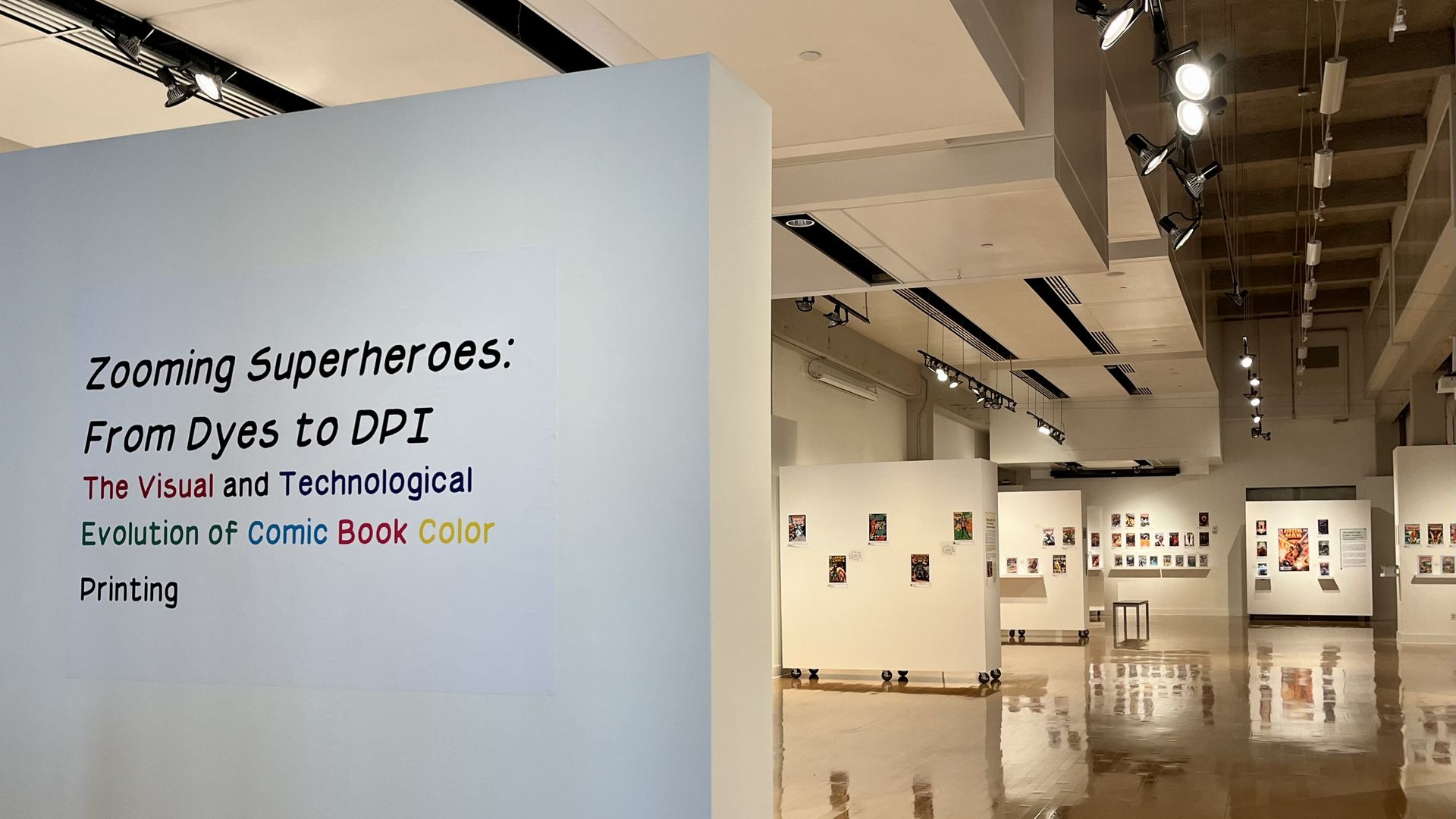 An art exhibit in the background and a white wall in the foreground that reads, "Zooming Superheroes: From Dyes to DPI, The Visual and Technological Evolution of Comic Book Color Printing"