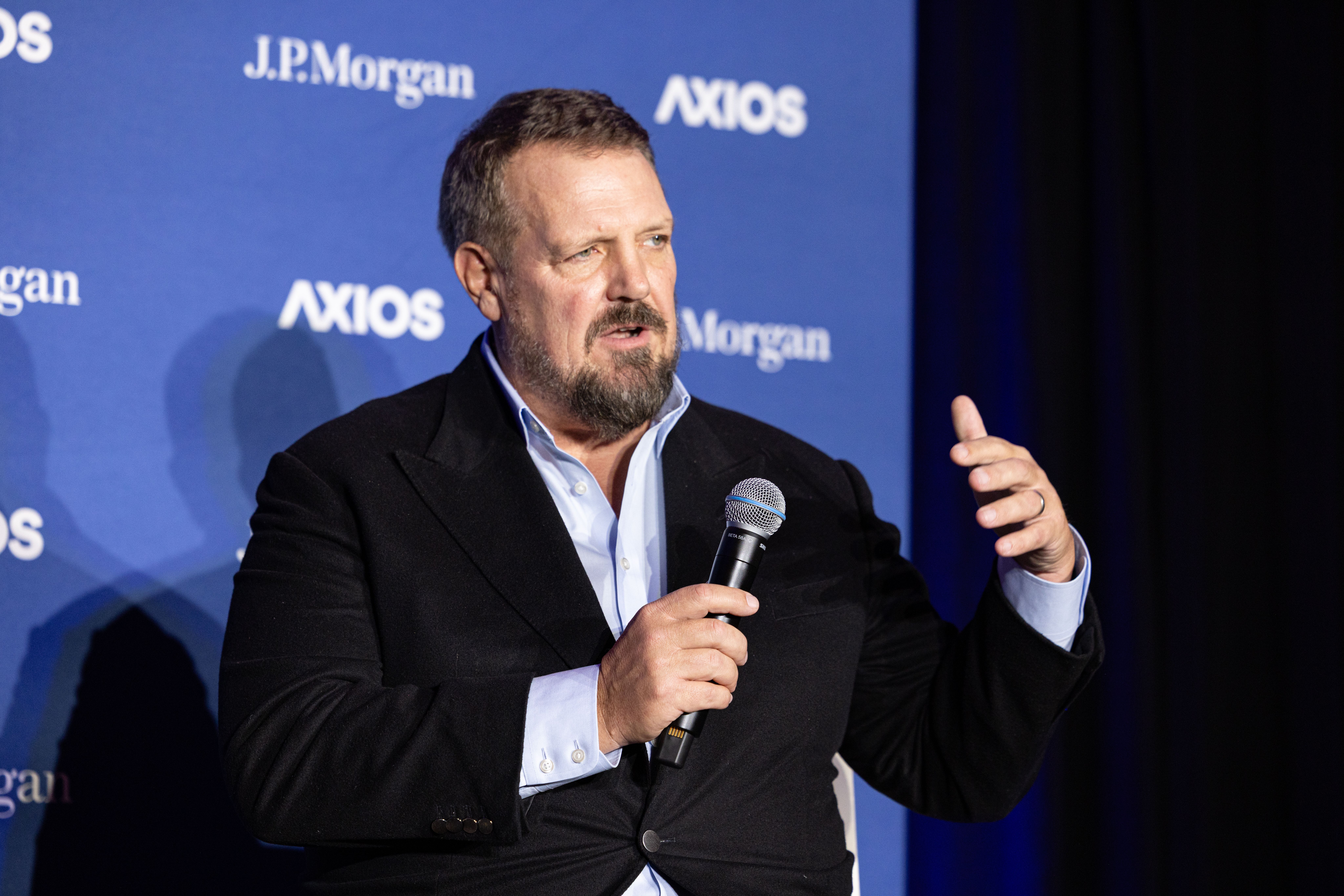 A man in a dark blazer and light blue shirt speaks into a handheld microphone on stage, gesturing with his right hand, against a blue backdrop showing "J.P. Morgan" and "AXIOS" logos.