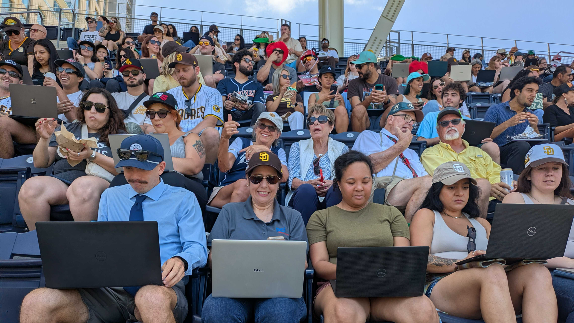 Crowded baseball stadium stands filled with fans, many in Padres gear, using laptops, tablets, and phones under a bright blue sky; a diverse crowd of ages and styles.