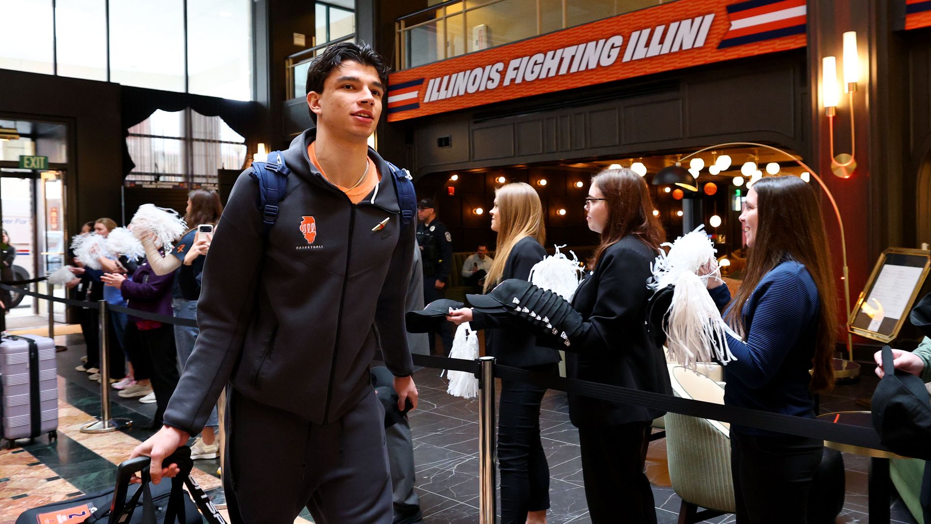 Young man in a dark tracksuit with an orange shirt and backpack pulls a black gym bag past cheerleaders with white pom-poms; an Illinois Fighting Illini banner hangs overhead in a busy lobby.