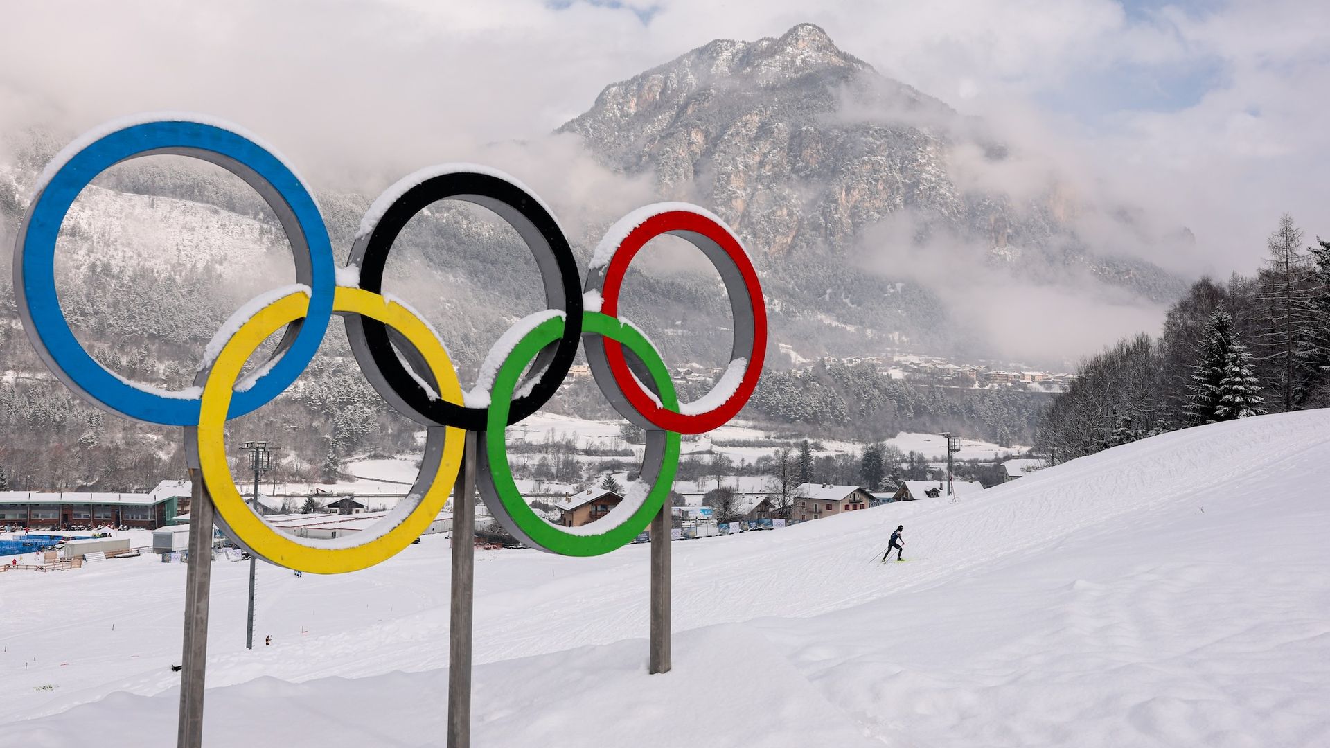 Blue, yellow, black, green and red Olympics rings statue in snow with a mountain in the background.