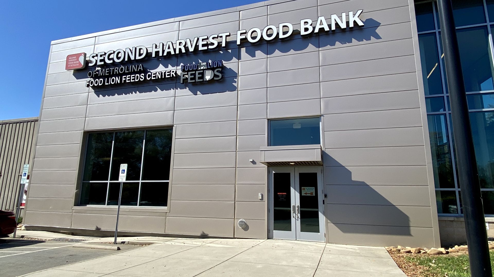Exterior of a modern building with silver panels displaying signs for Second Harvest Food Bank of Metrolina and Food Lion Feeds Center, under a clear blue sky.