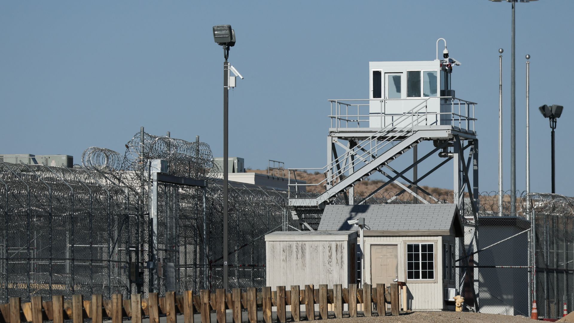 Exterior view of a prison facility with tall chain-link fences topped with barbed wire, a guard tower, multiple security cameras, and wooden barriers in front under a clear blue sky.