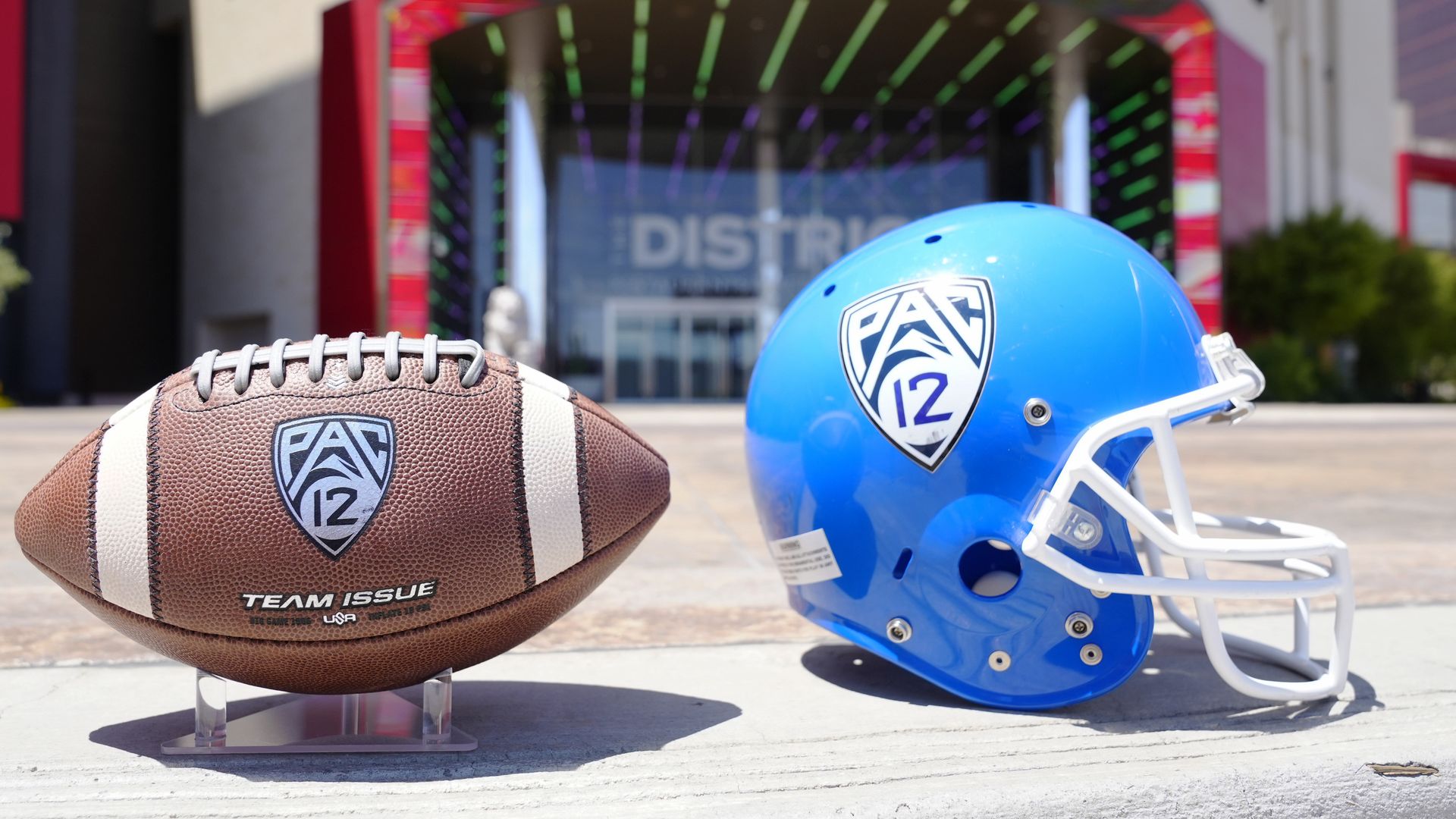 A football and a football helmet with Pac 12 logos on them on the ground in front of the entrance to a stadium.