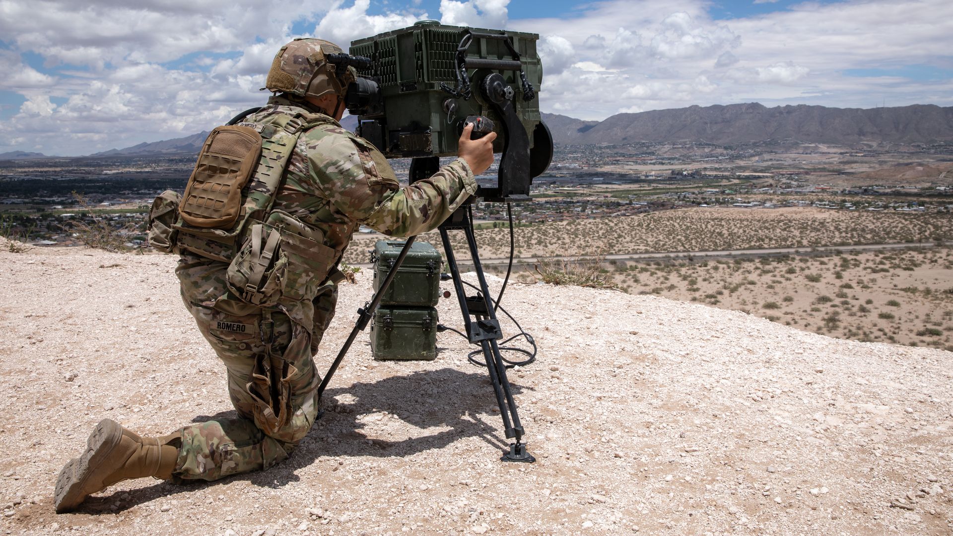 Soldier in camouflage uniform kneels on dusty terrain operating a large tripod-mounted military device with a desert city and mountain range in the background under a partly cloudy sky.