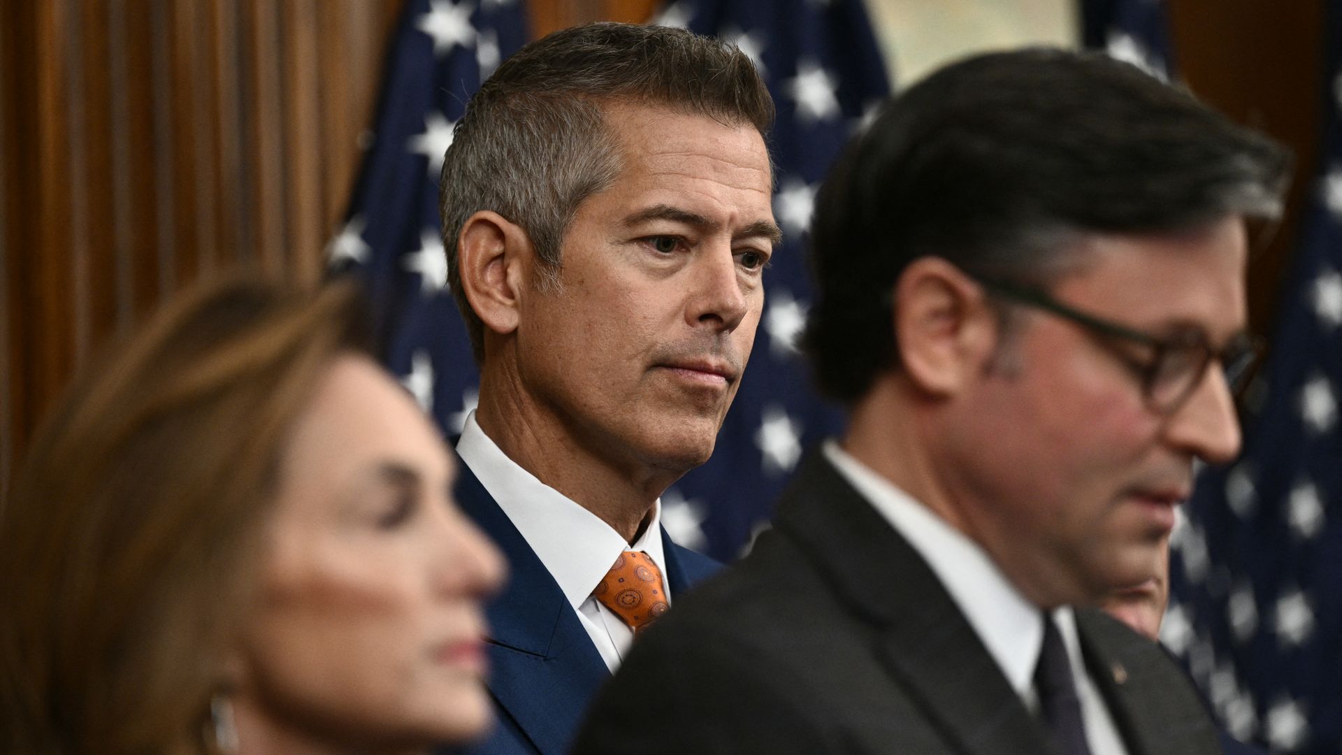 Sean Duffy — wearing a dark suit, white shirt and orange tie — looks down as Mike Johnson, wearing a dark suit and glasses, speaks in the foreground.