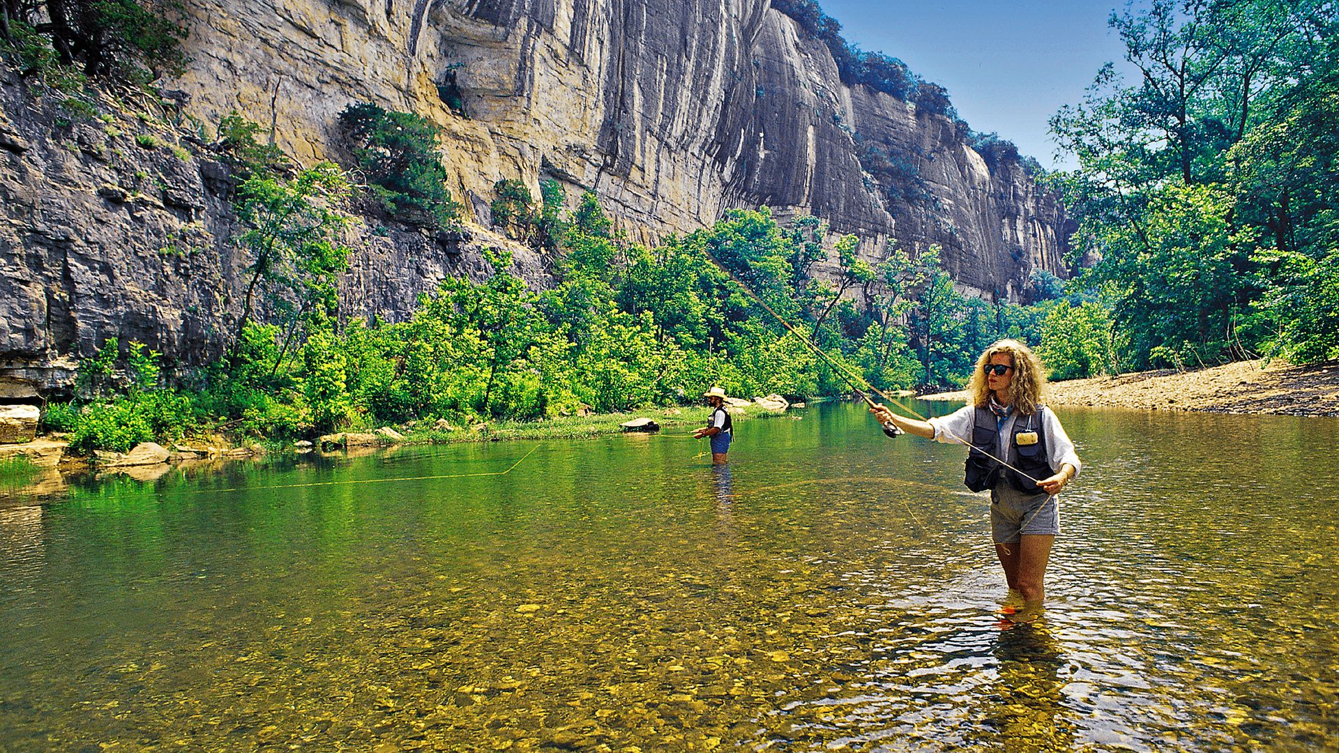 A photo of two people standing in a river fly fishing with bluffs in the background. 