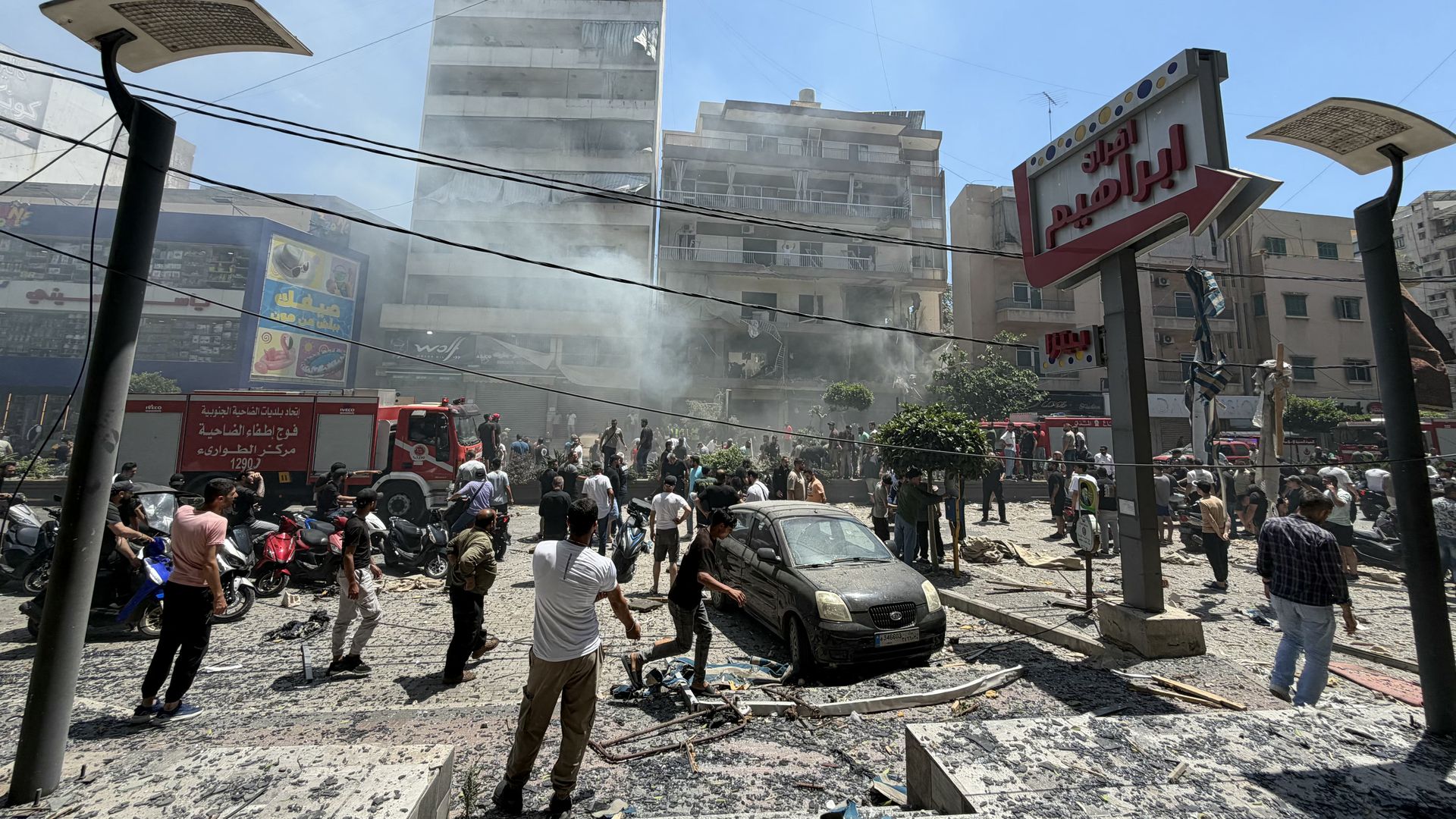 A crowd stands outside a damaged building in Beirut’s southern suburbs after an Israeli airstrike, with people surveying the scene and debris visible nearby.