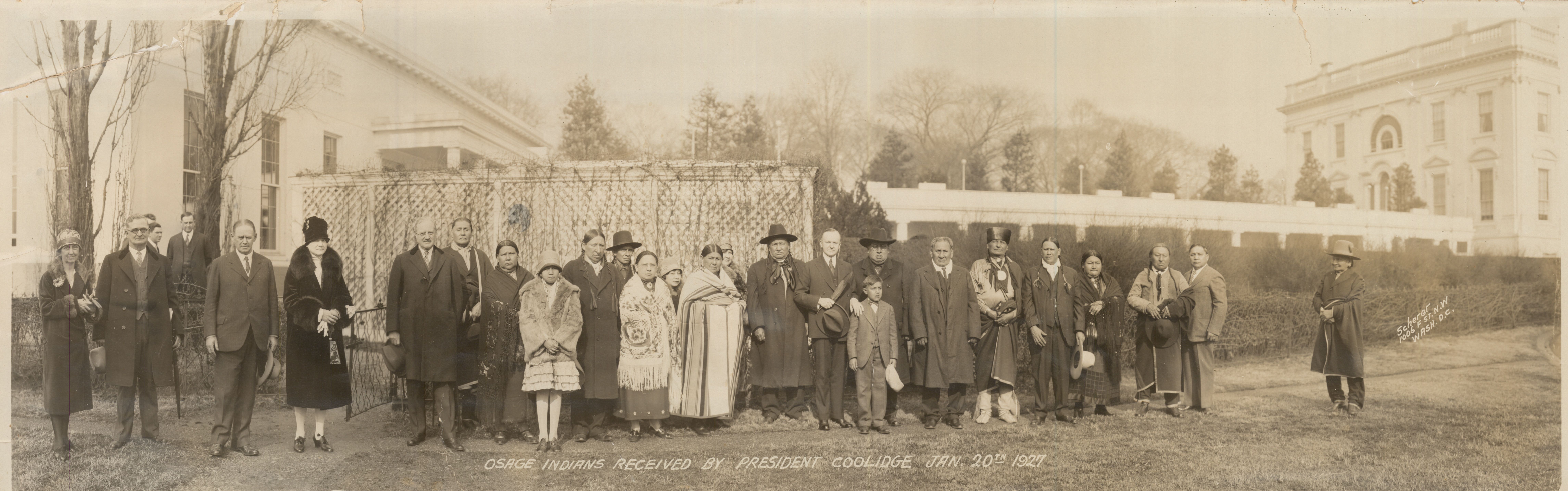 President Calvin Coolidge (center right, with hat in hand) and unidentified representatives from the Osage Nation stand in front of the East Colonnade at the White House in 1927, with trees in the background in this sepia photo.
