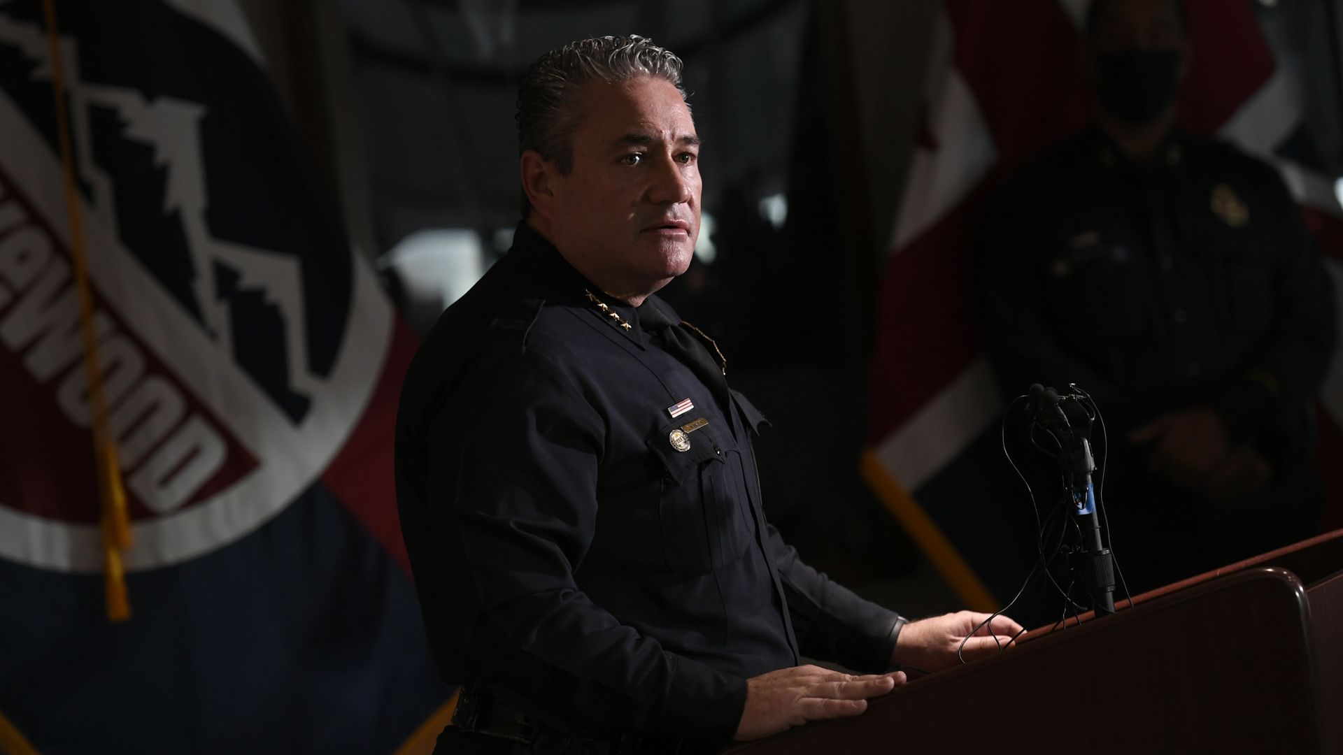 A police chief in full uniform stands near a lectern in a dimly-lit room while addressing member of the press.  