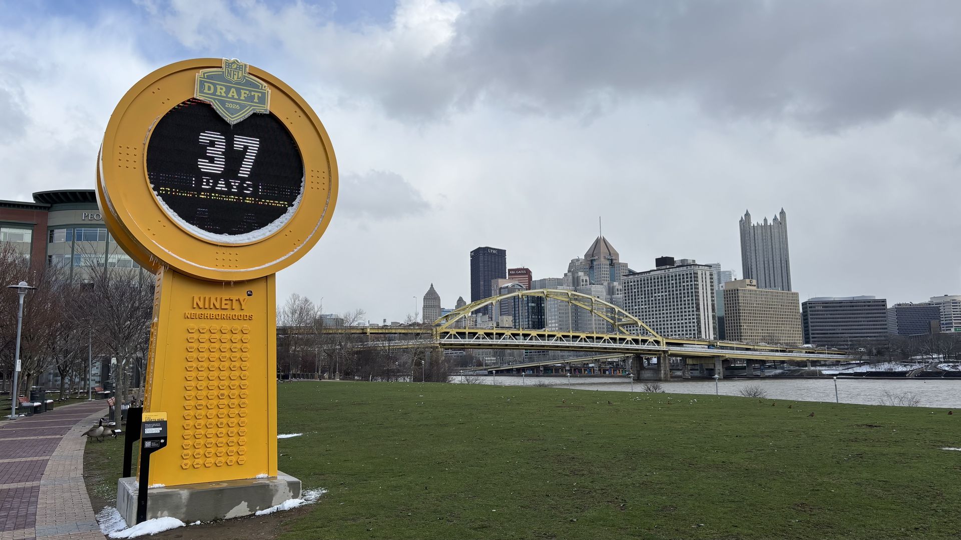 Large yellow NFL Draft sign with a digital 37 on display sits in a riverfront park; a curved yellow arch bridge spans the river, with Pittsburgh's skyline rising under a cloudy sky.