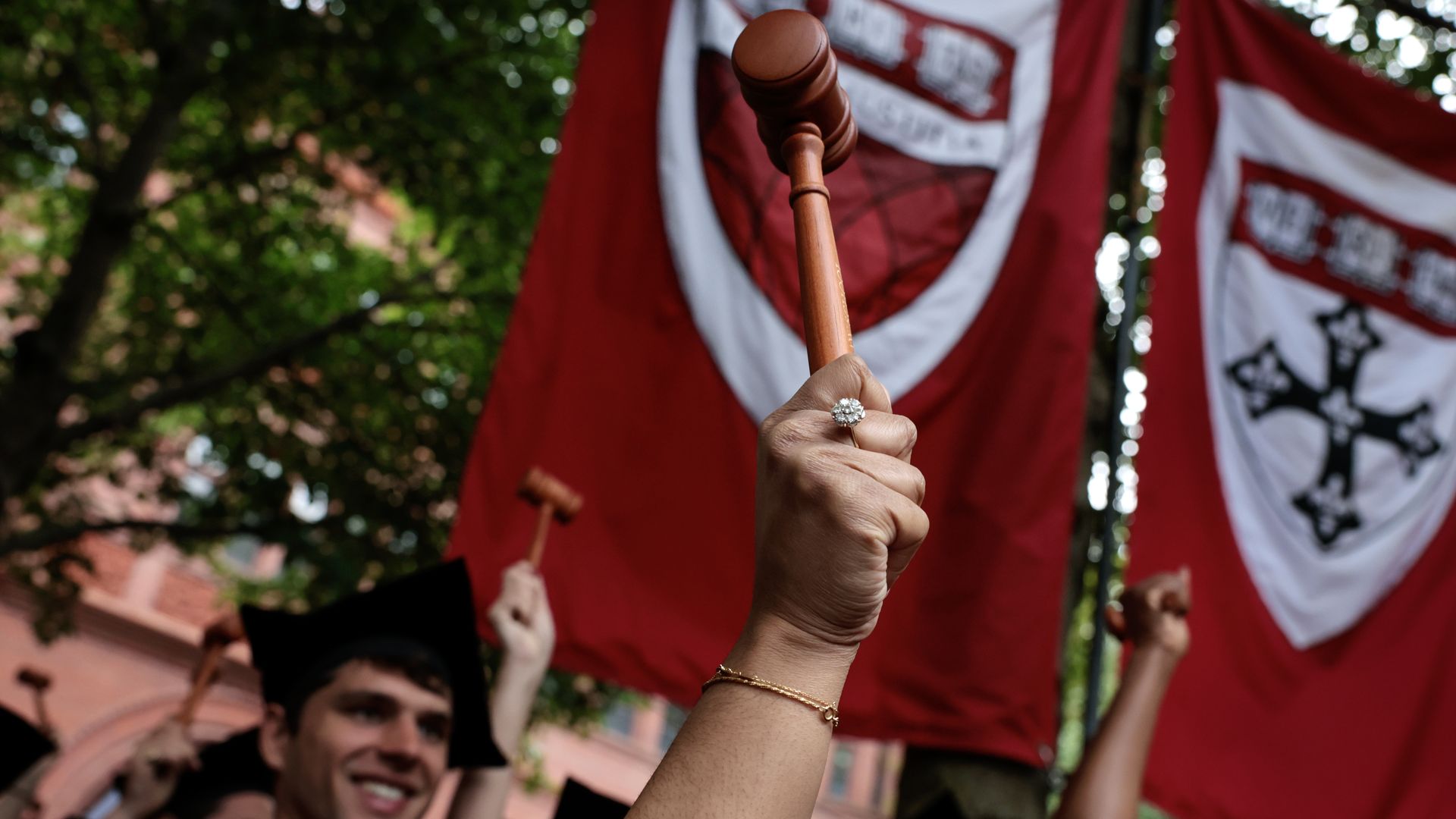 Graduates in black caps and gowns hold wooden gavels high, with a close-up of a hand wearing a diamond ring and gold bracelet, red banners and green trees in the background.