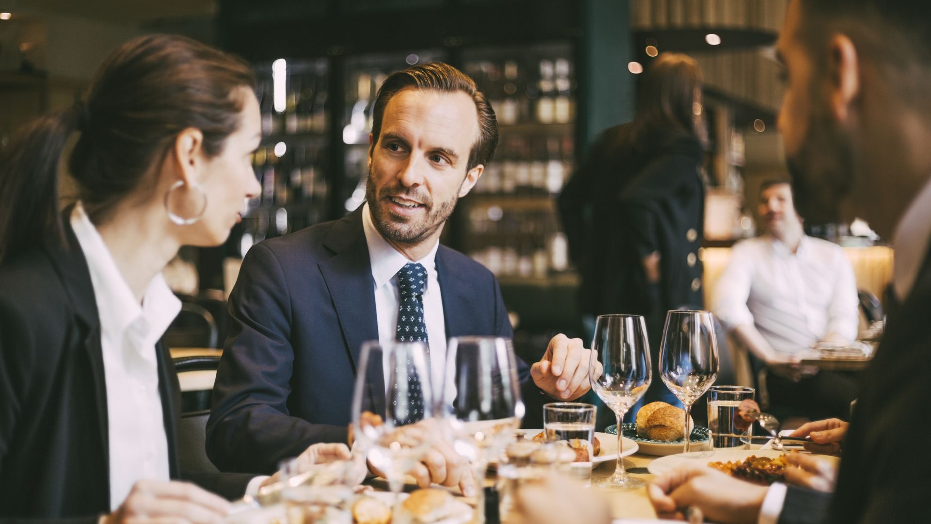 Group of professionals in a restaurant; center man in a navy suit and dotted tie chats with a woman on his left, others mingle in the background, with wine glasses and plates on the table.