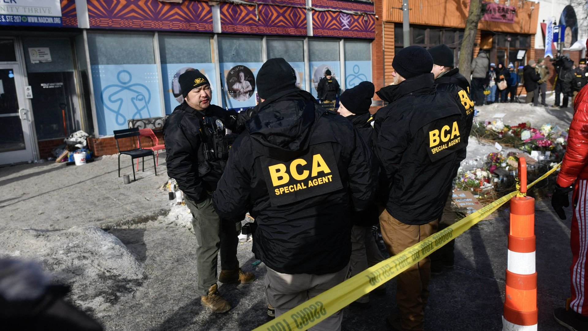 Minnesota Bureau of Criminal Apprehension special agents wearing black jackets, standing near yellow police tape in front of a building with purple and orange patterns; people and floral tributes visible nearby on a snowy day.