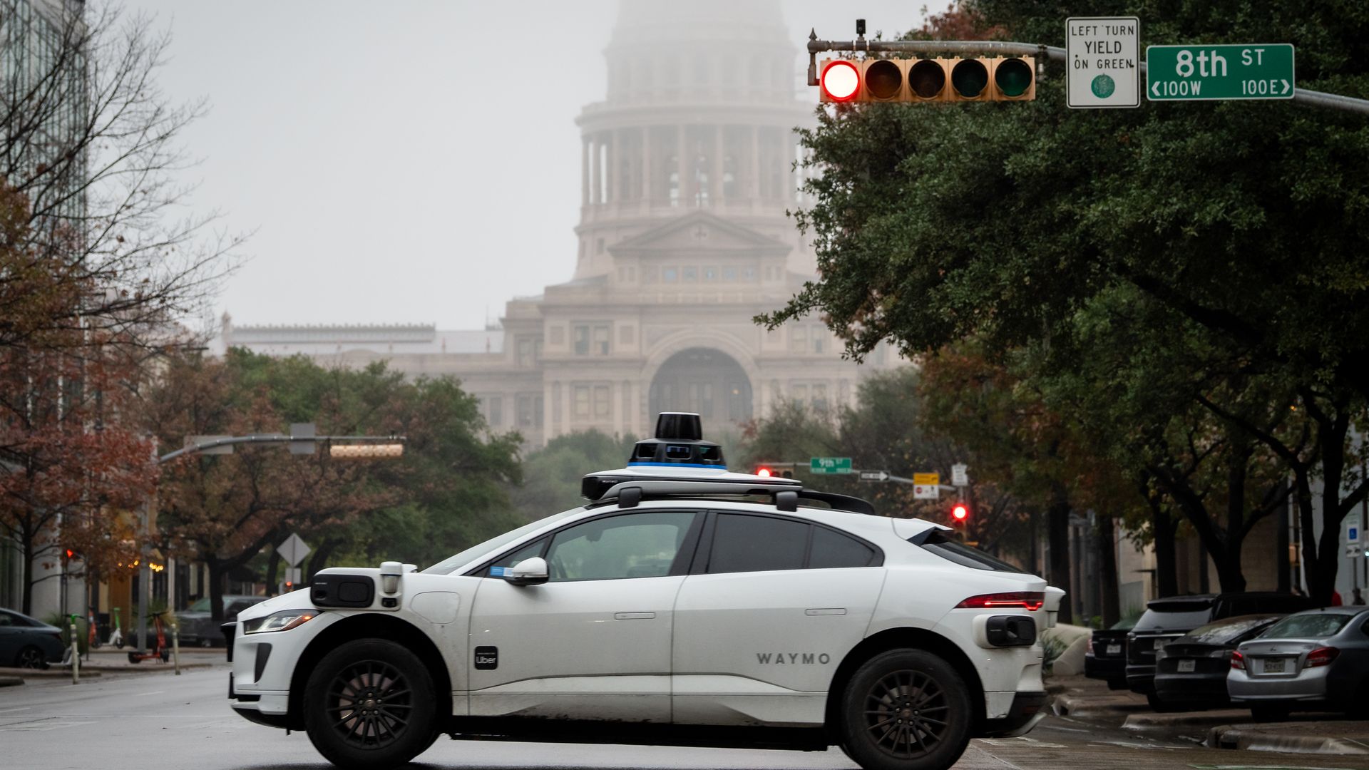 White Waymo self-driving car with Uber logo stopped at red light on 8th St in front of a large dome building on a foggy day, surrounded by trees and parked cars.