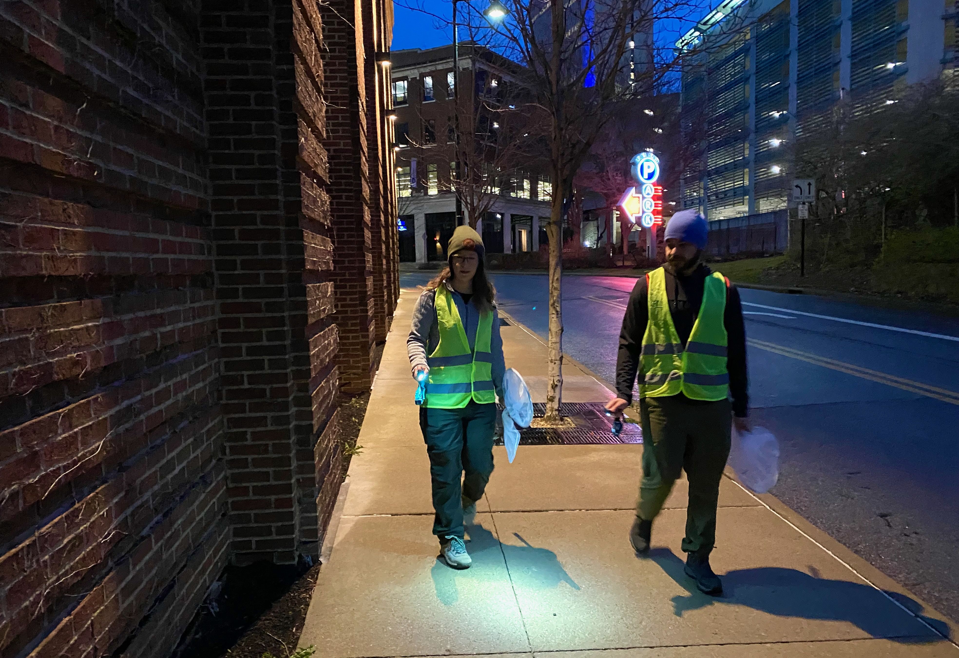 A man and woman in neon yellow vests walk down a street near a brick wall, carrying nets and flashlights